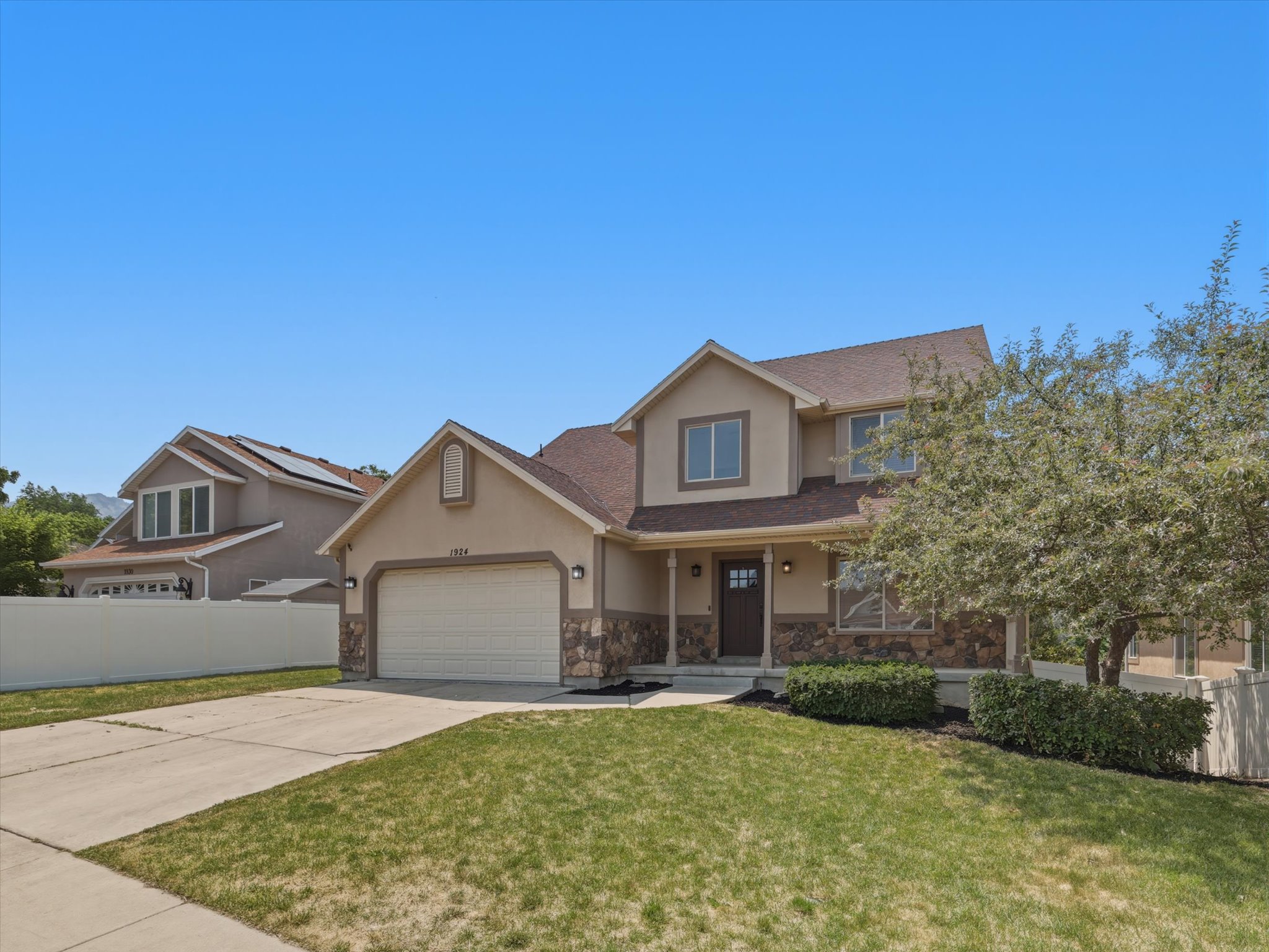 Two-story residence featuring a stone and stucco exterior, an attached garage, a covered front porch, and mature landscaping