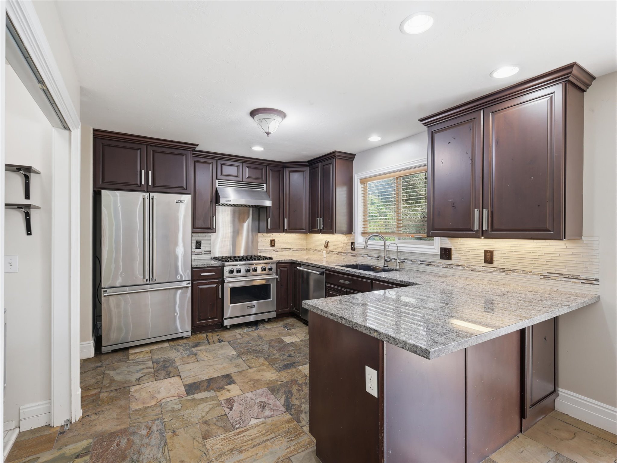 Kitchen featuring dark wood cabinetry, stainless steel appliances, stone tile flooring, granite countertops, and a mosaic tile backsplash