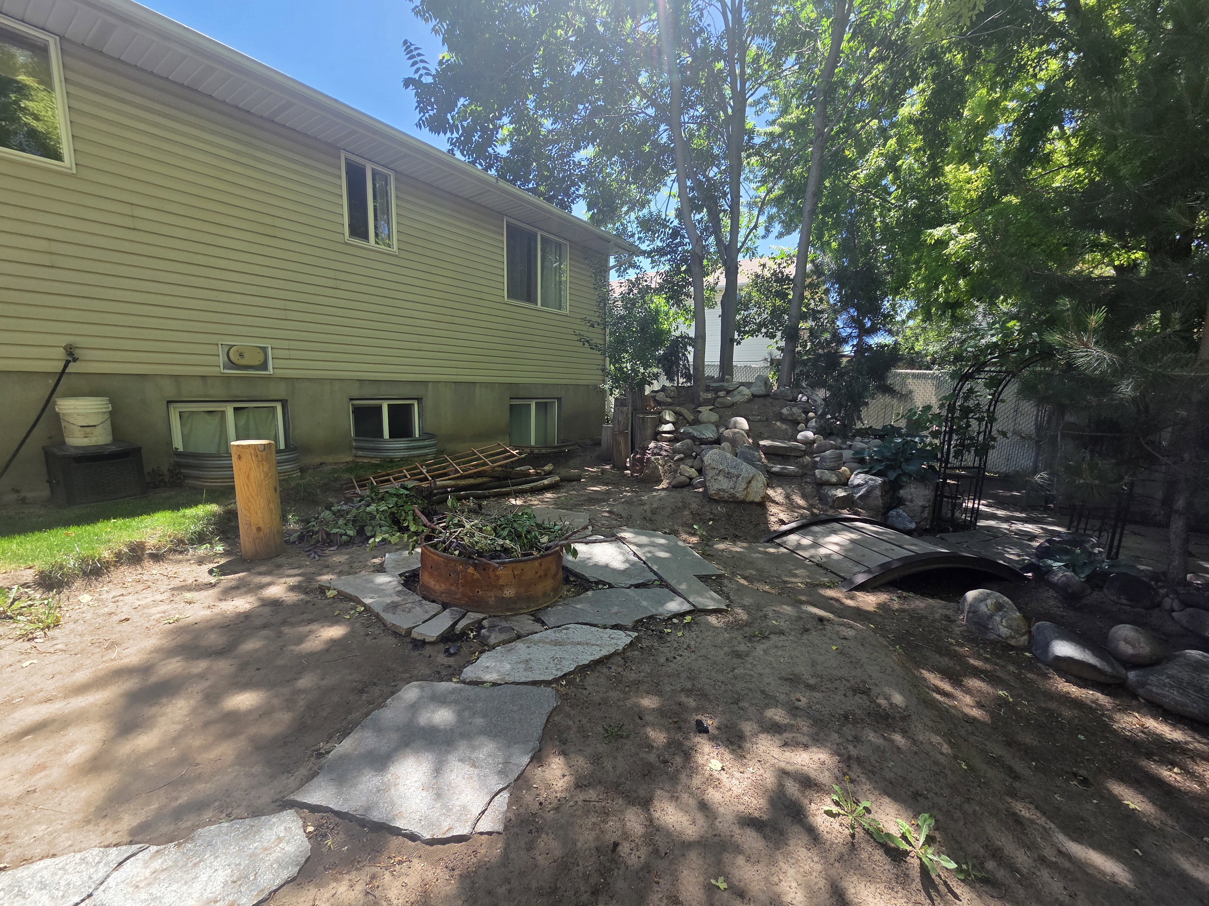 Shaded backyard featuring mature trees, a stone pathway, a decorative wooden bridge, and a rock garden