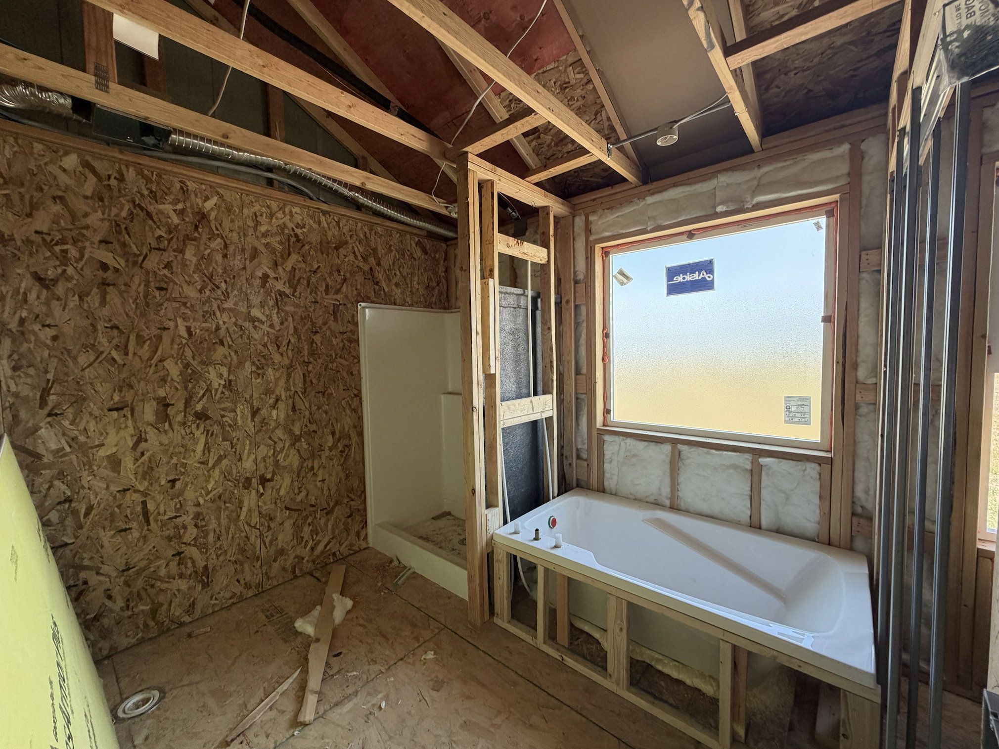Bathroom space under construction featuring a built-in bathtub, a separate shower stall, a window, exposed wood framing, and oriented strand board walls