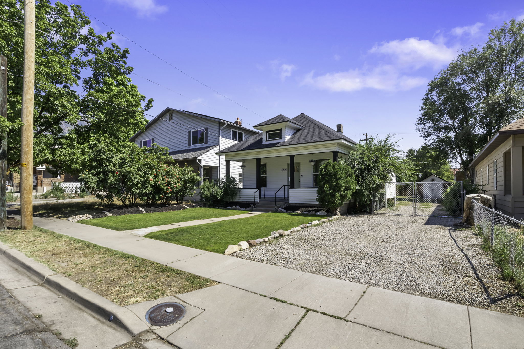 Classic white residence featuring a covered front porch with dark pillars, a gravel driveway, a grassy front yard, and mature landscaping