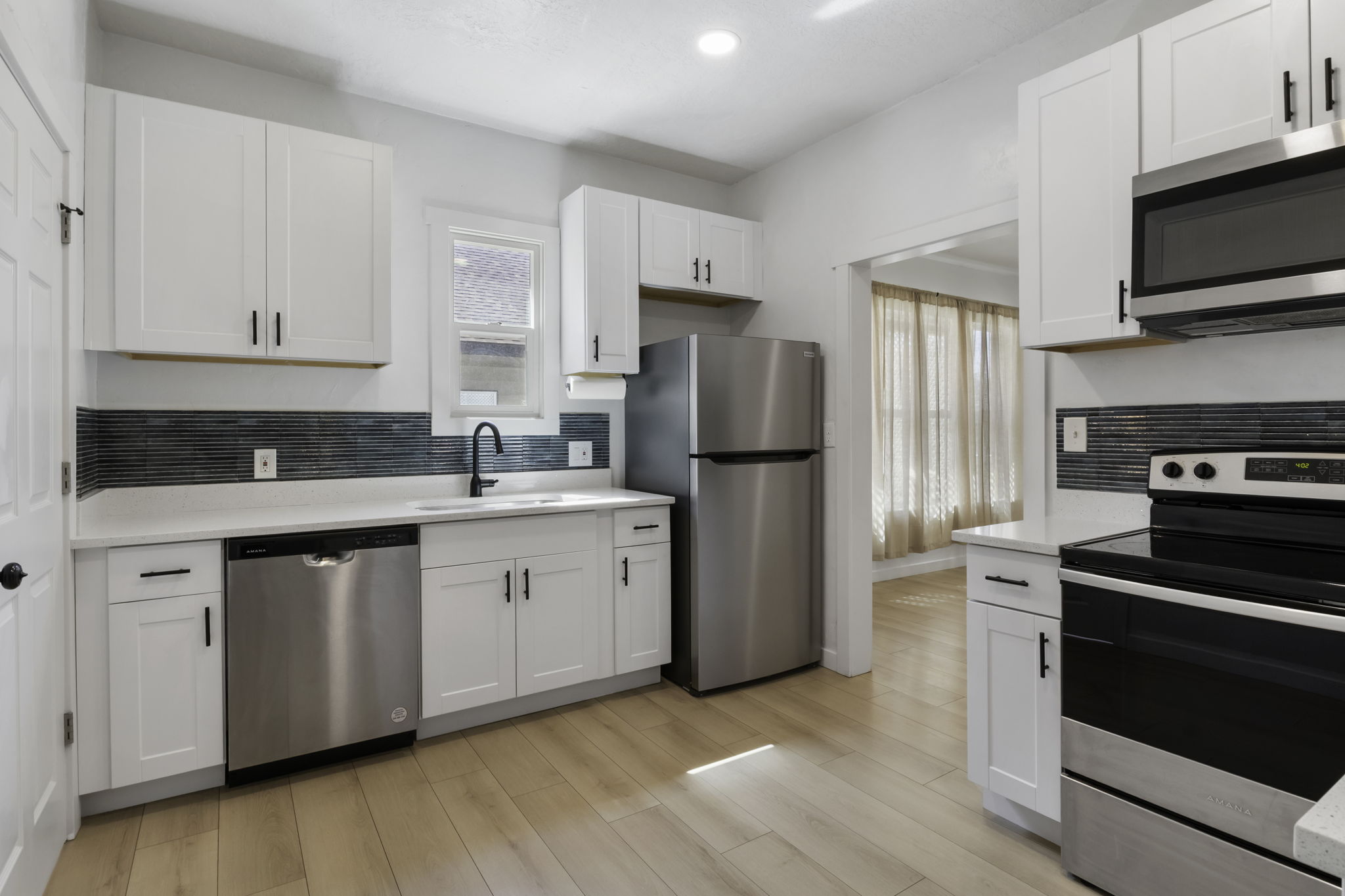 Modern kitchen featuring light wood-finish flooring, white shaker cabinetry with matte black hardware, stainless steel appliances, a dark linear tile backsplash, and white countertops