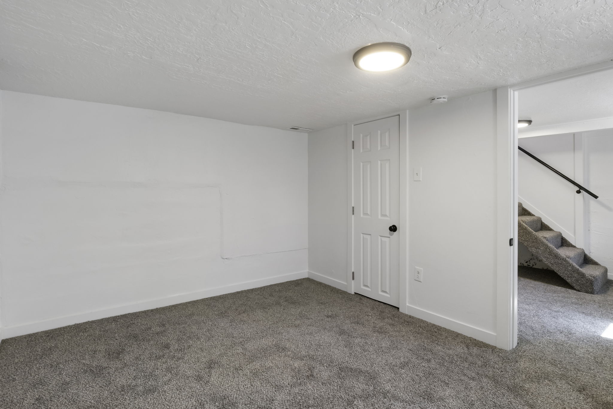 Carpeted room featuring a white paneled door with a black handle, a flush-mount ceiling light, and access to a carpeted staircase with a black handrail