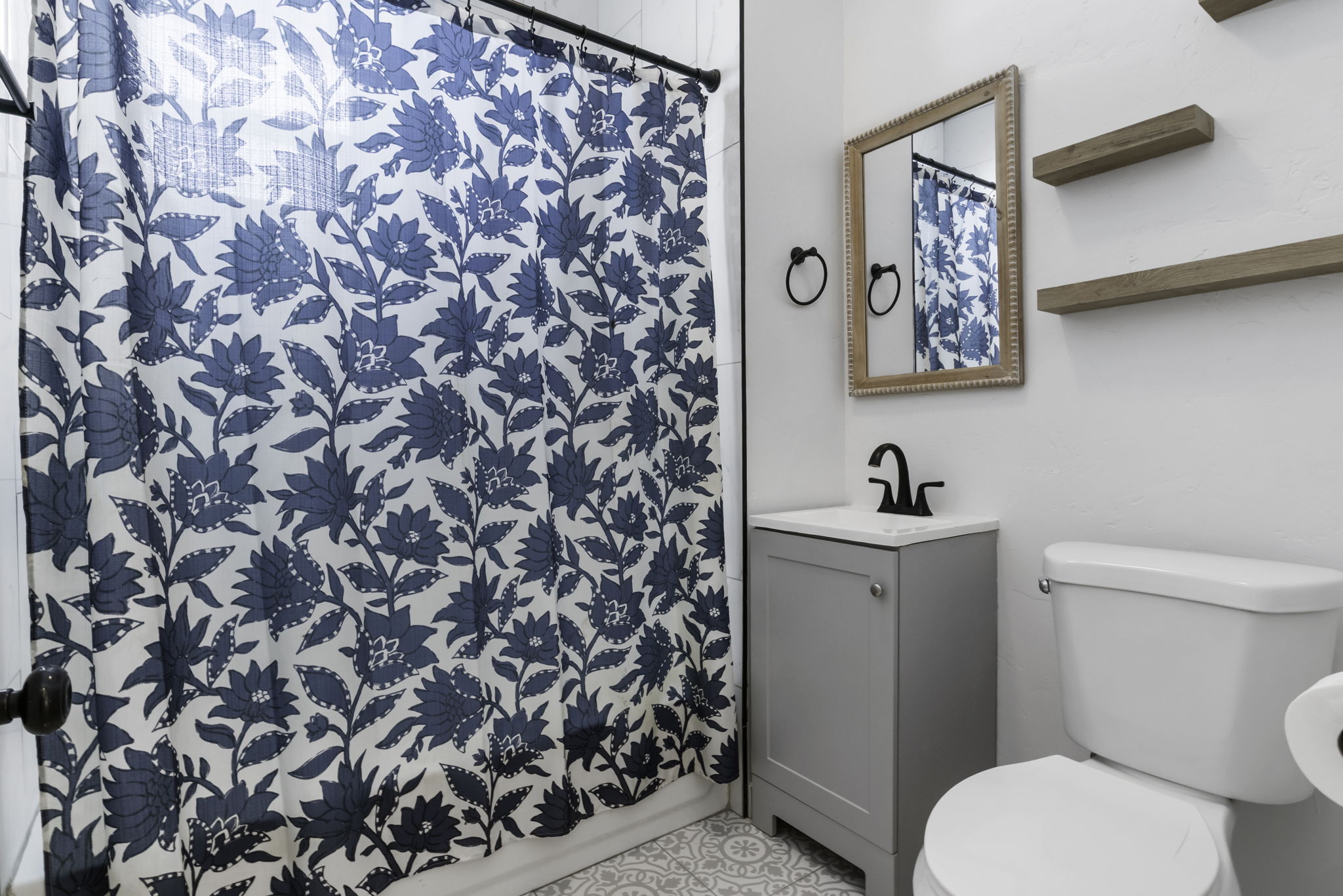 Bathroom featuring a vanity with a light gray cabinet and white countertop, black matte faucet, a framed mirror, and two wooden floating shelves