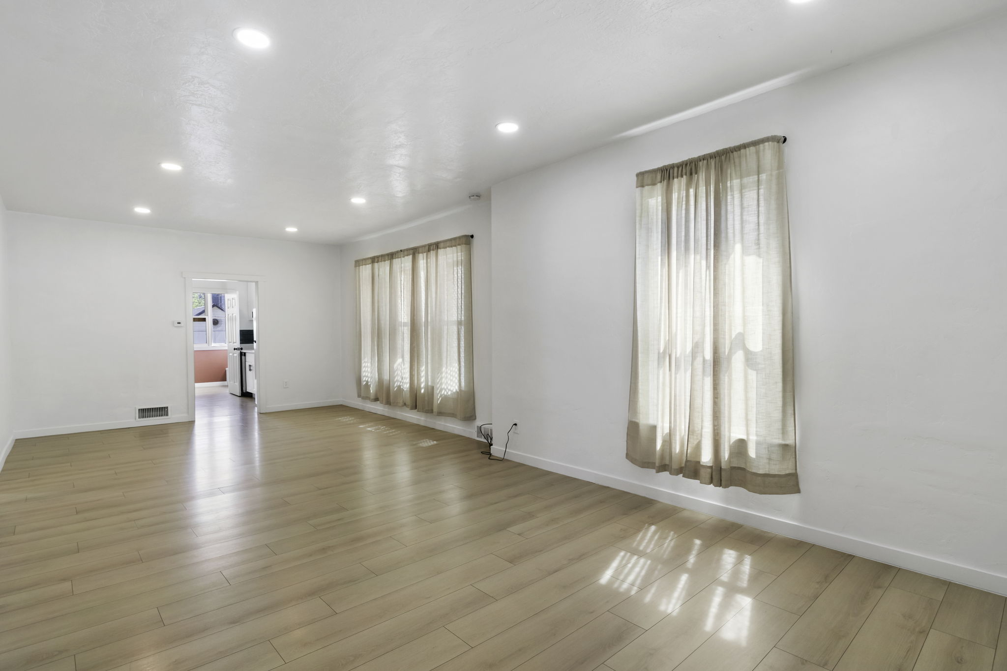 Spacious living area featuring light wood-finish flooring, recessed lighting, and multiple windows with sheer drapery