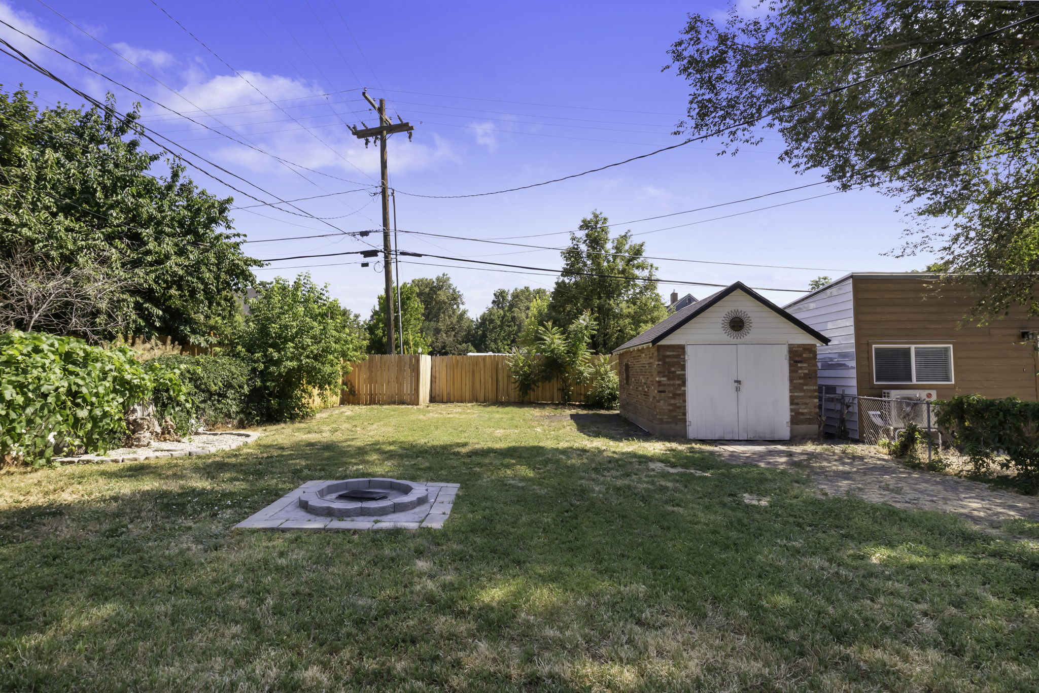 Expansive grassy backyard featuring a built-in fire pit with stone surround, a detached brick garage with white doors, wood fencing, and mature perimeter trees