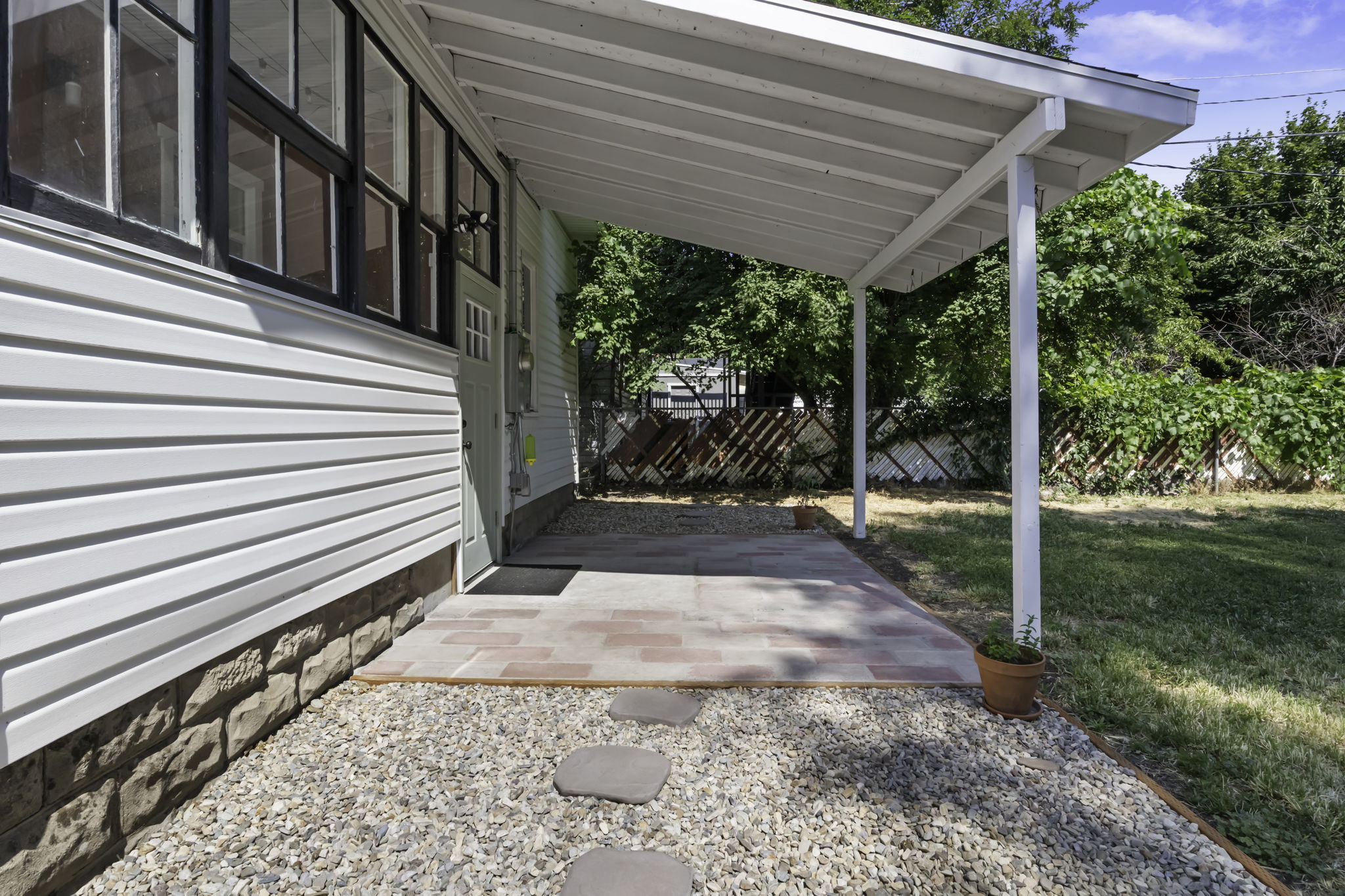 Covered patio with brick paver flooring and gravel landscaping, featuring a white vinyl siding exterior with a stone veneer foundation