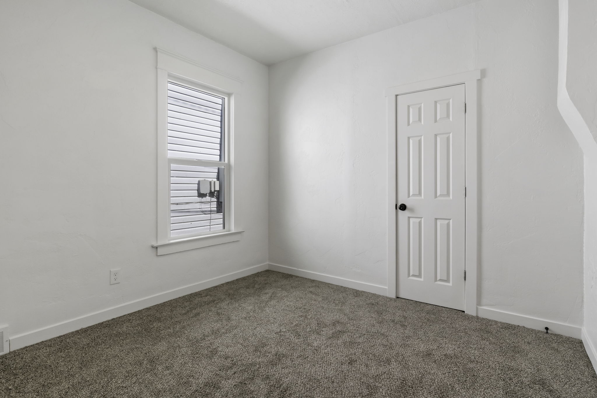 Room featuring a window with white blinds, a six-panel door with a black knob, light-colored walls, and textured carpeting