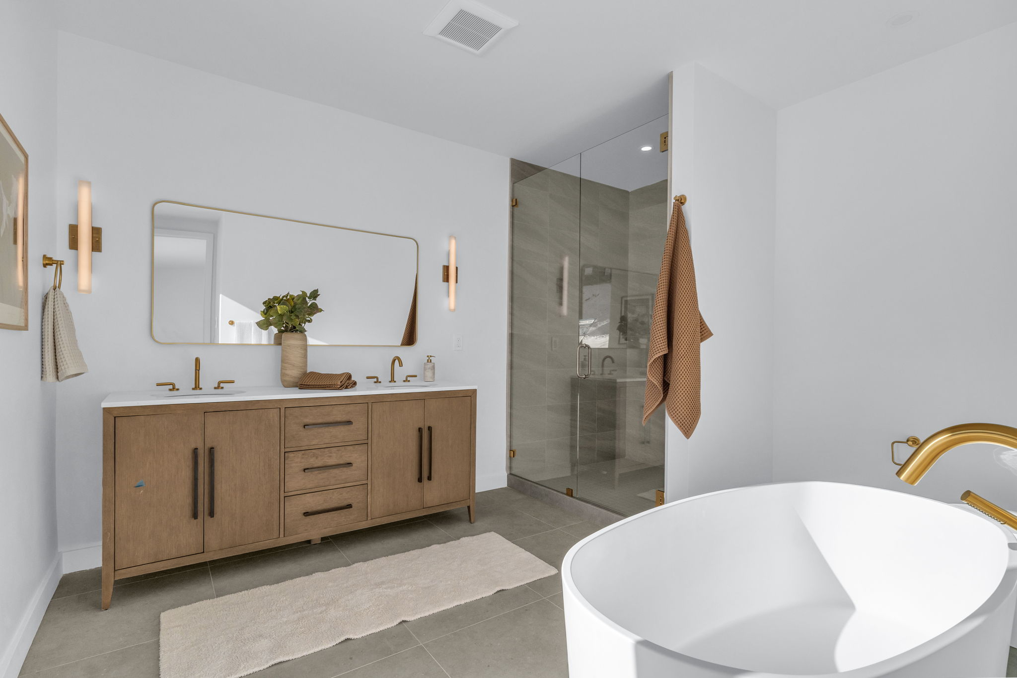 Spacious bathroom featuring a dual vanity with a white countertop and wood-finish cabinetry, a freestanding soaking tub with gold-tone fixtures, a glass-enclosed shower with tiled walls, and rectangular wall-mounted sconces