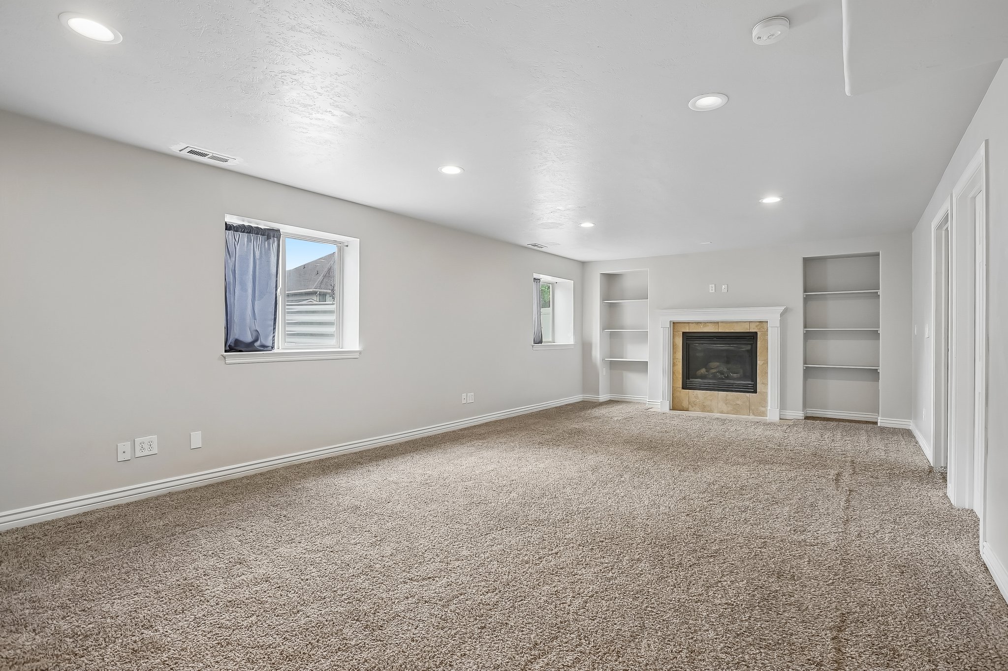 Spacious room featuring neutral carpeting, a fireplace with a light-colored surround, two built-in shelving units, recessed lighting, and two windows