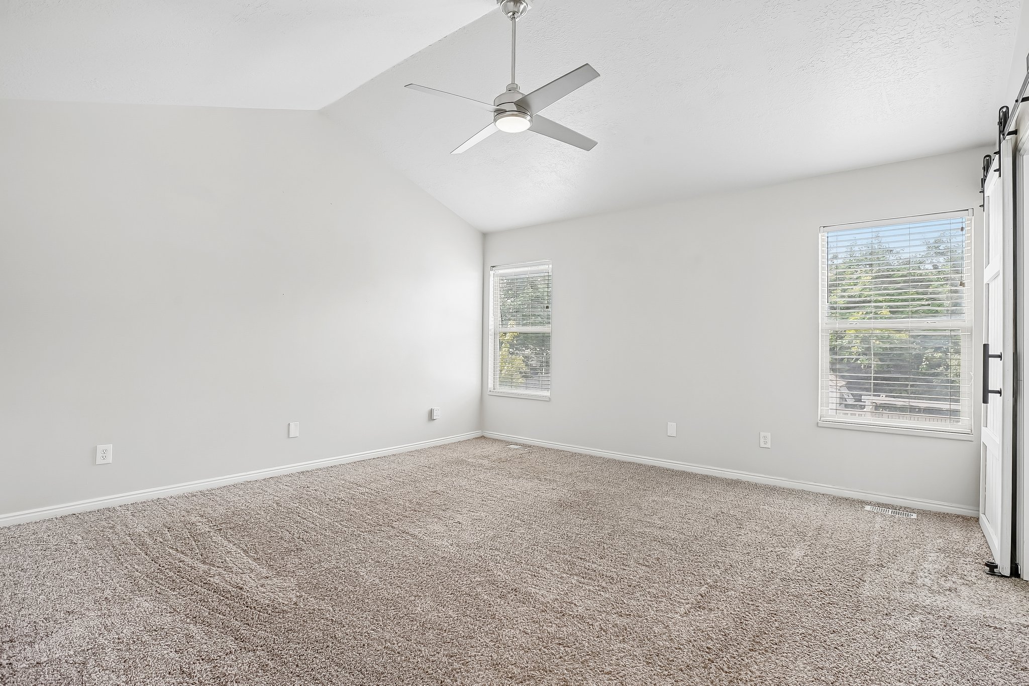 Spacious room featuring vaulted ceilings, light-colored carpeting, two windows with blinds, a ceiling fan, and a barn door with black hardware
