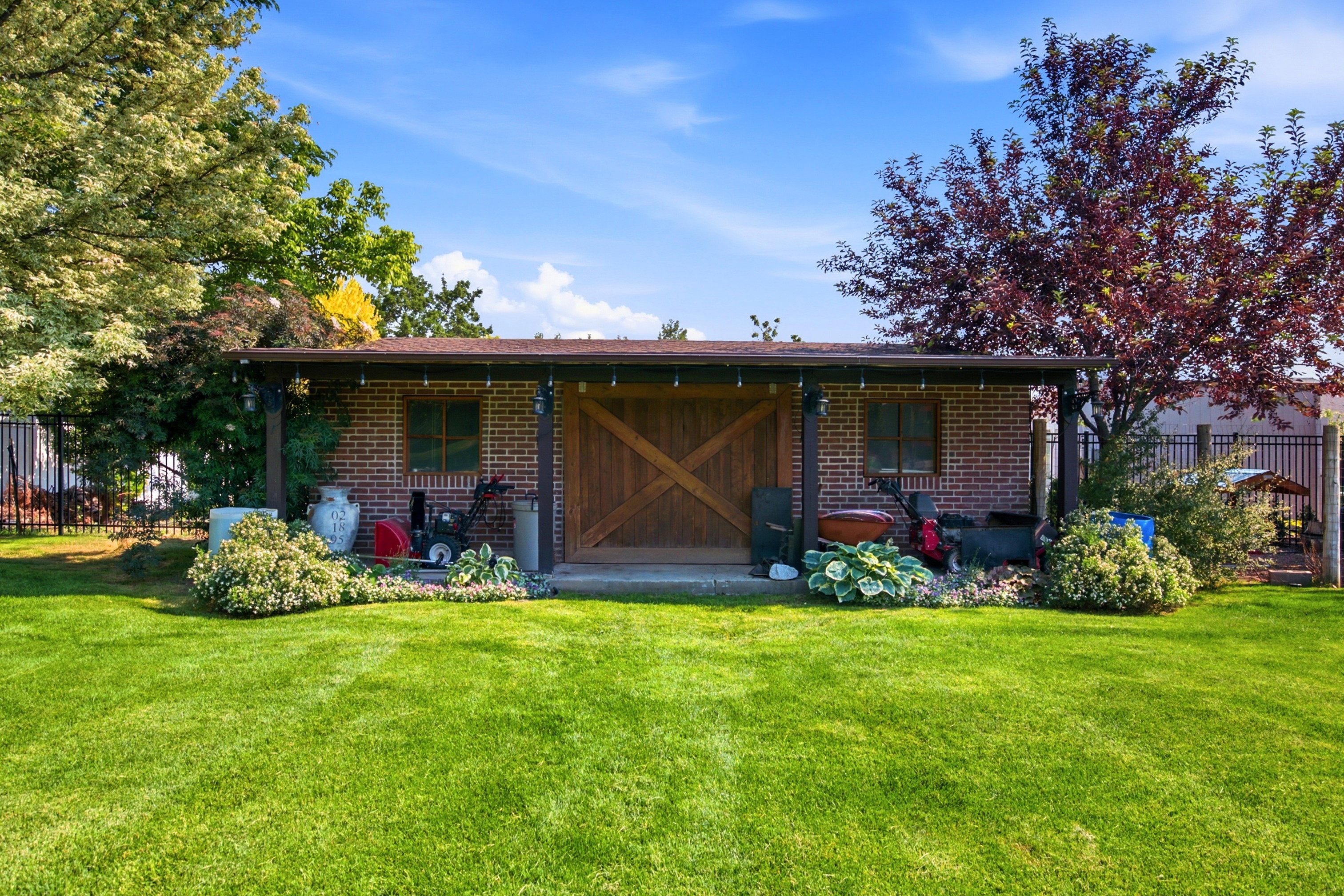 Expansive green lawn with a brick outbuilding featuring a covered porch, wooden cross-buck doors, and a window