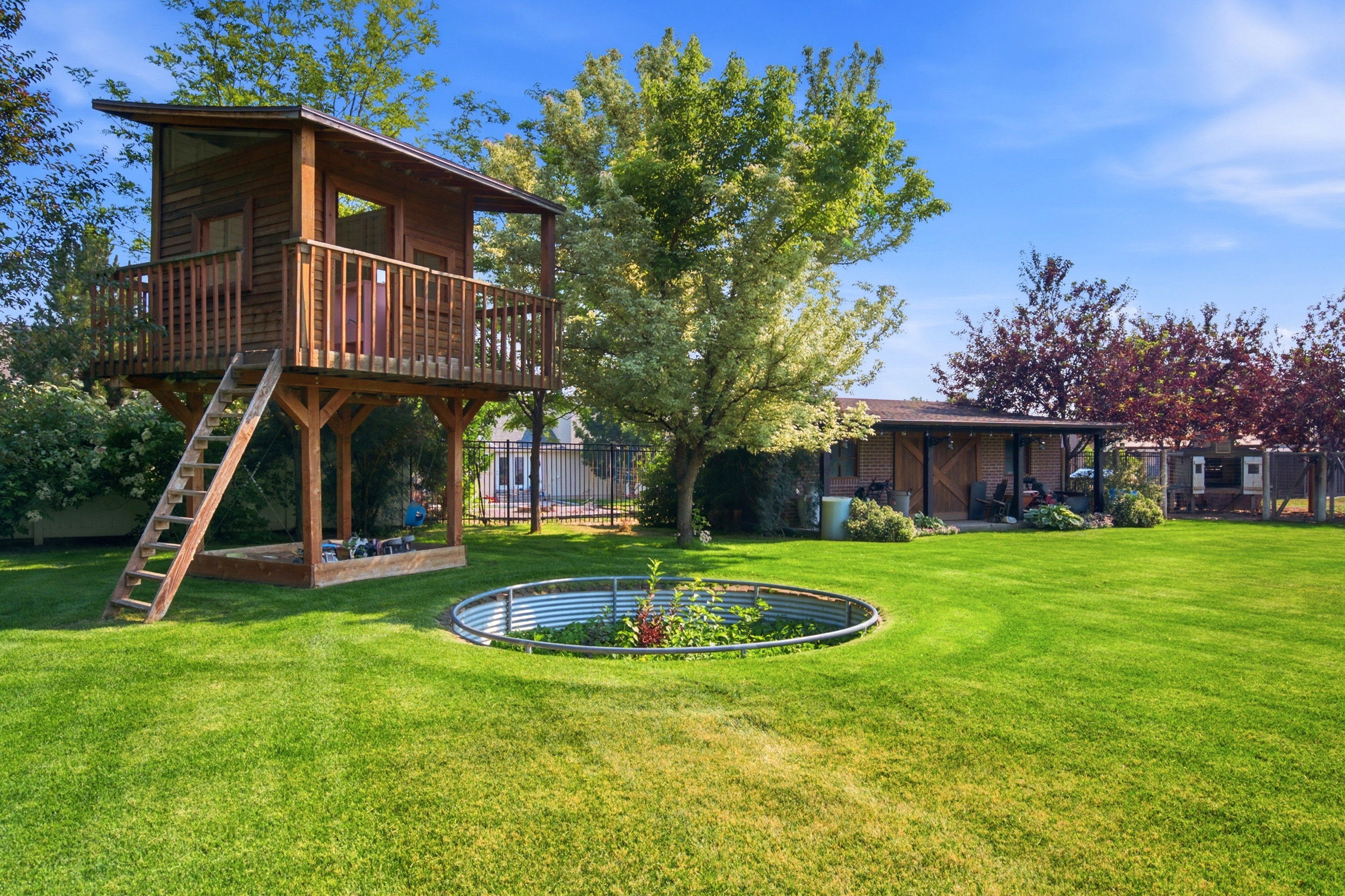 Expansive backyard featuring a custom wooden treehouse with a balcony, a circular raised garden bed, a covered patio area with string lighting, and mature trees