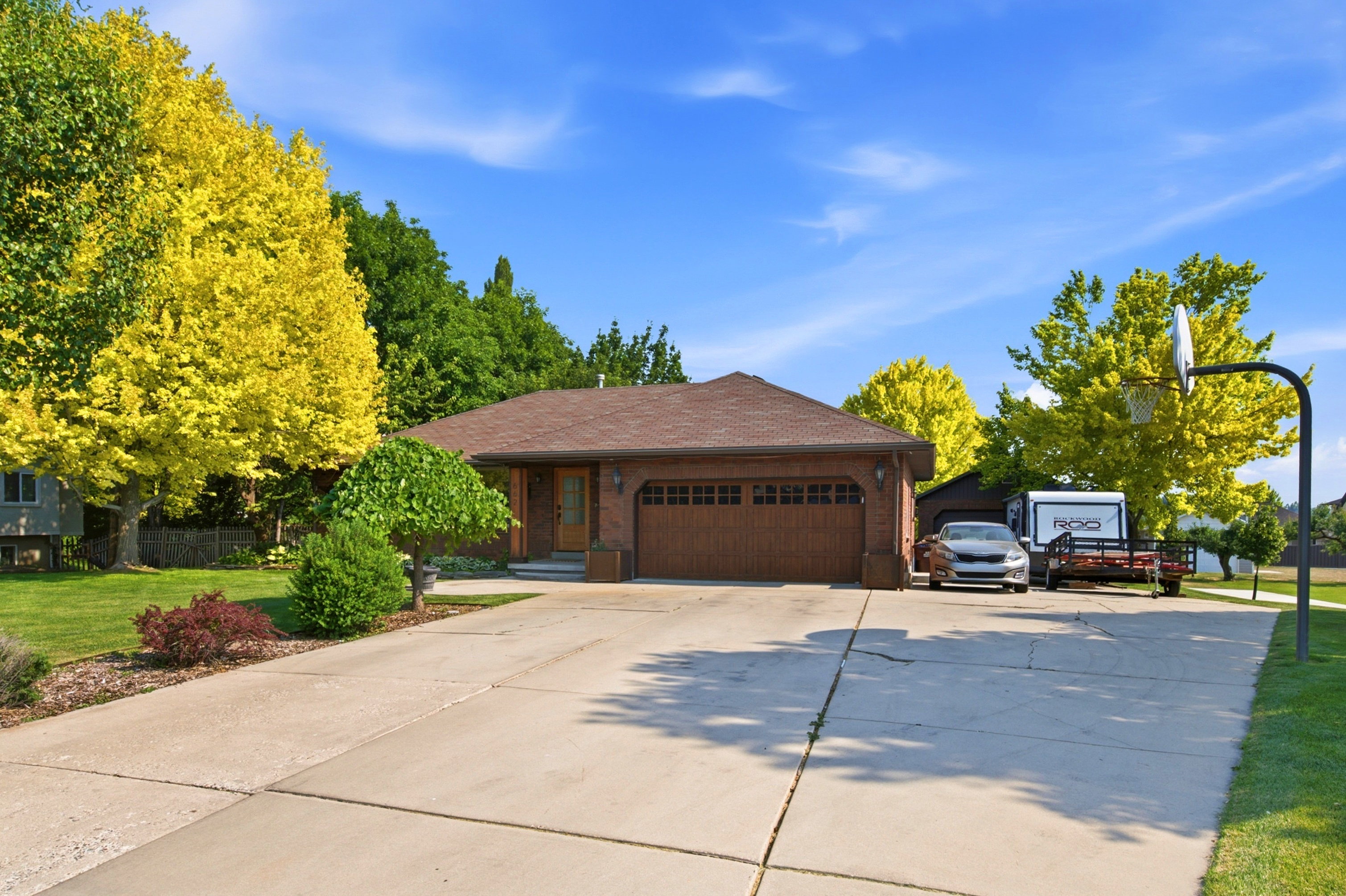 Brick facade residence featuring a brown shingle roof, a two-car garage with a wood-finish door, a concrete driveway, and mature landscaping with vibrant trees