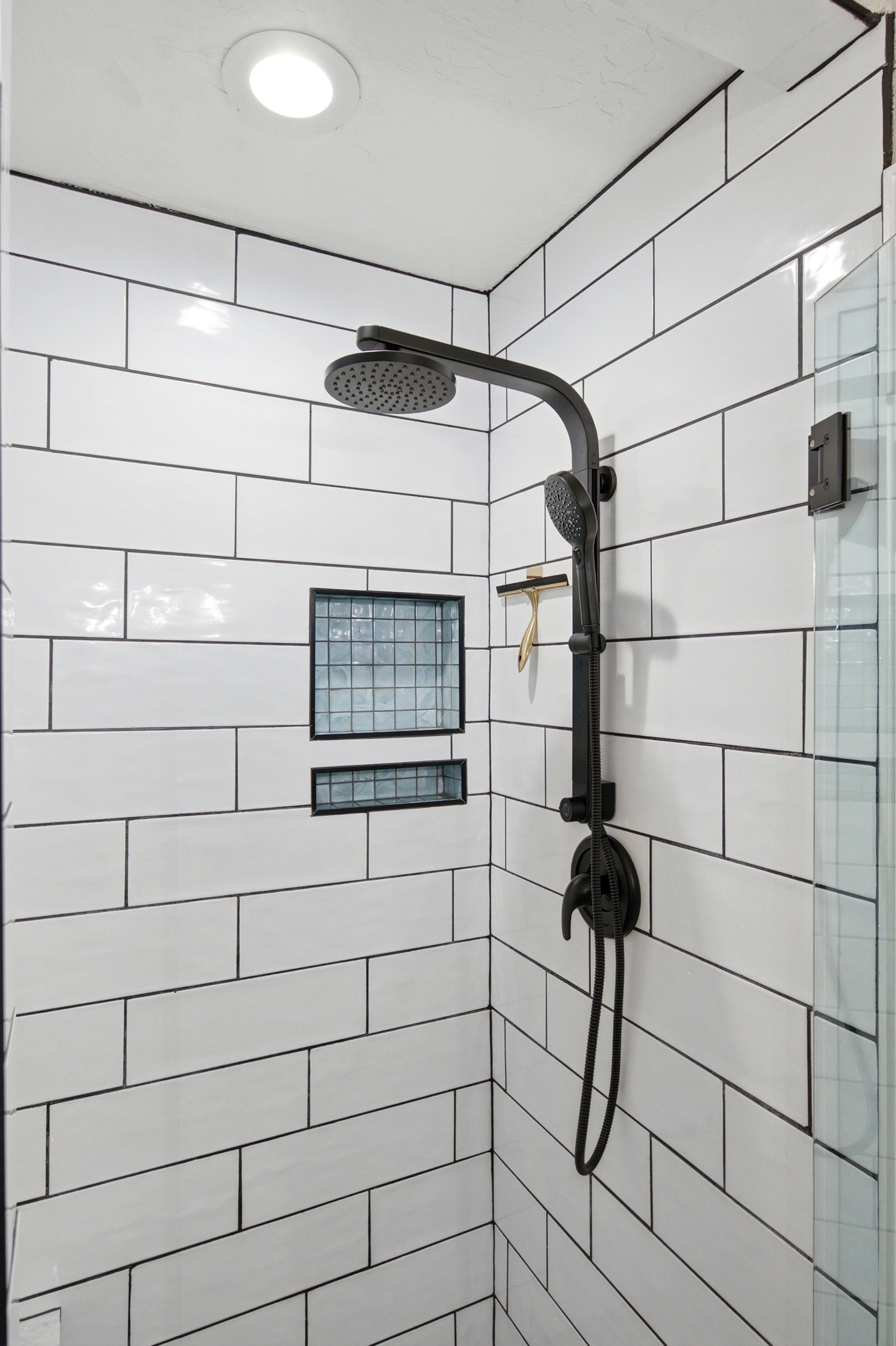 Shower stall featuring white subway tile with dark grout, a matte black dual shower head system, and recessed shelving with glass block accents
