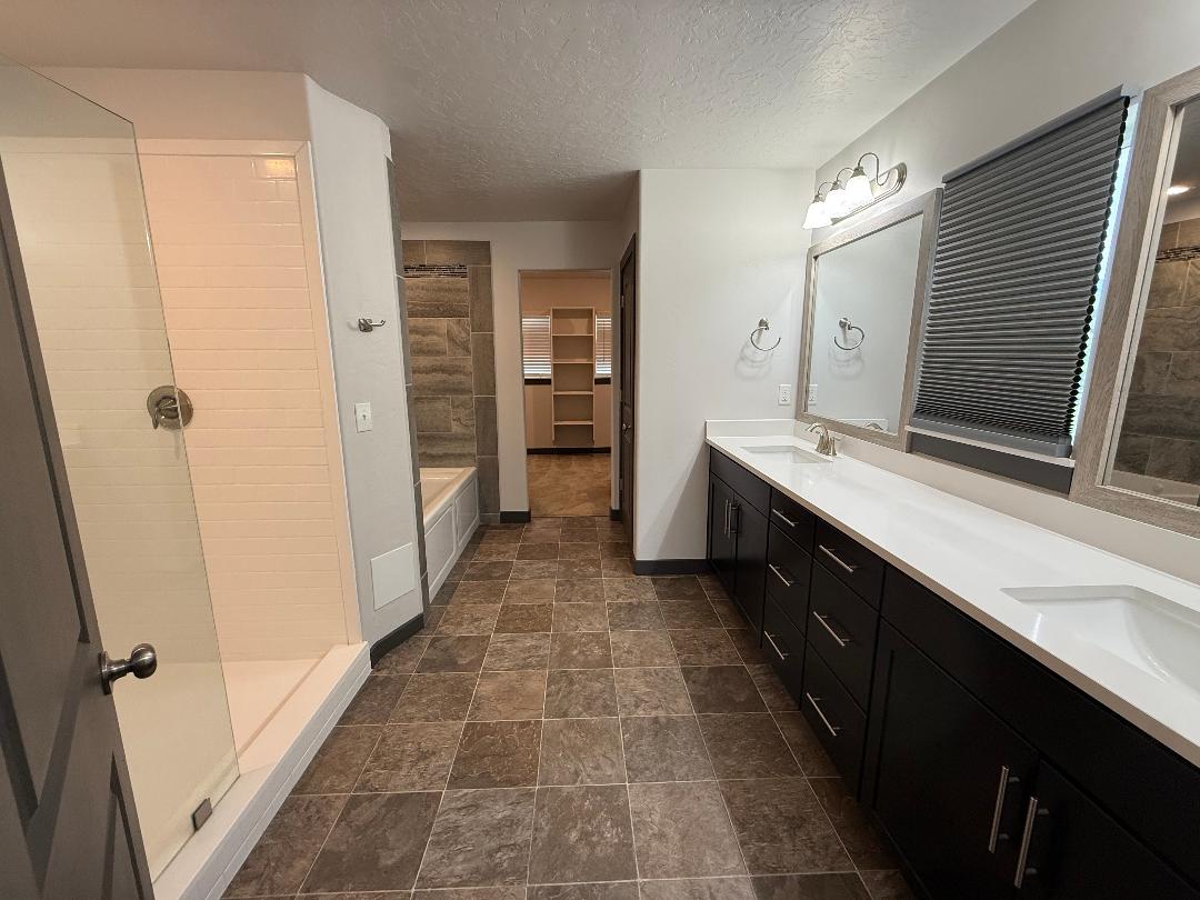 Bathroom featuring a dual vanity with white countertops and dark cabinetry, a glass-enclosed shower, a built-in bathtub, and tiled flooring