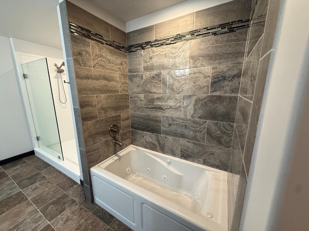 Master bathroom featuring a white jetted tub, a separate glass-enclosed walk-in-shower with white surround  and large format wall and floor tiles with a decorative mosaic accent band