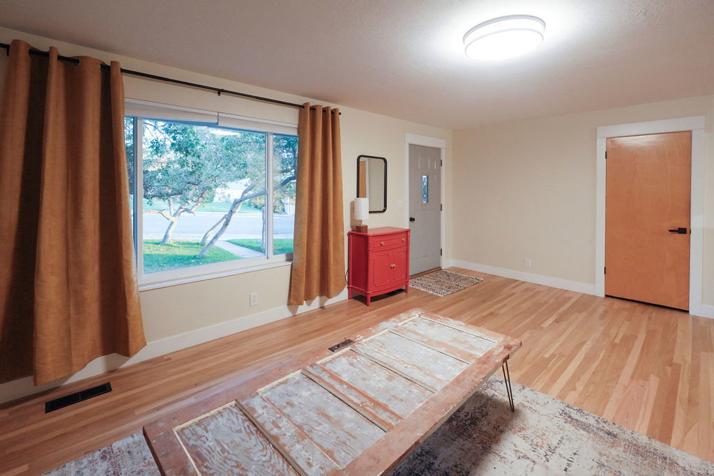 Bright living area featuring wood-finish flooring, a large window, a ceiling-mounted light fixture, a light-colored entry door with a decorative glass panel, and a solid wood interior door with black hardware