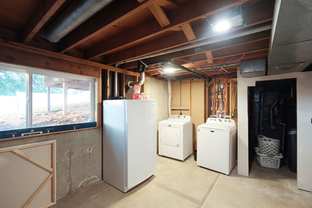 Concrete floor basement with exposed wood joists and utility lines, featuring a laundry area with washer and dryer, a window, and a built-in water heater