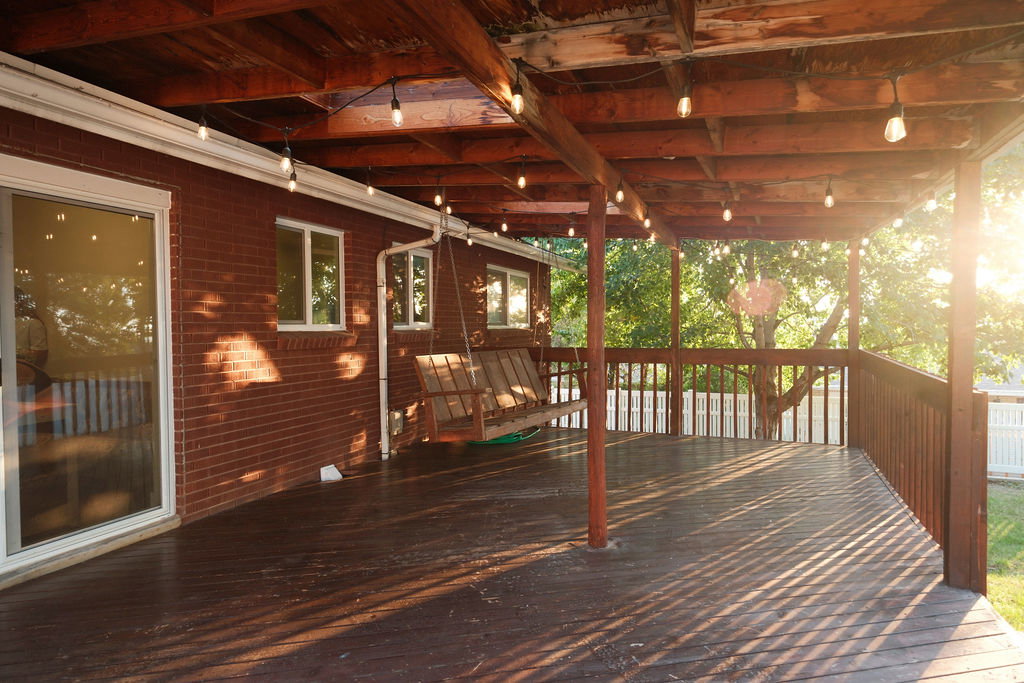 Expansive covered wood deck featuring a rich stain finish, exposed wood beam ceiling, decorative string lighting, and a white perimeter fence