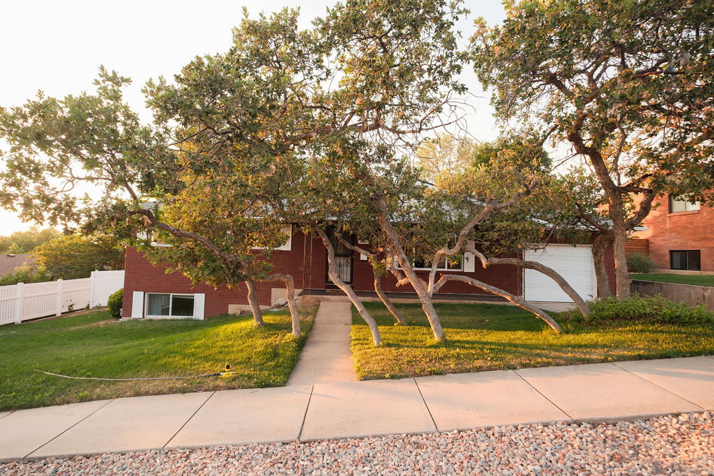 Mature trees frame the red brick exterior, featuring a white garage door, concrete walkway, and a white picket fence