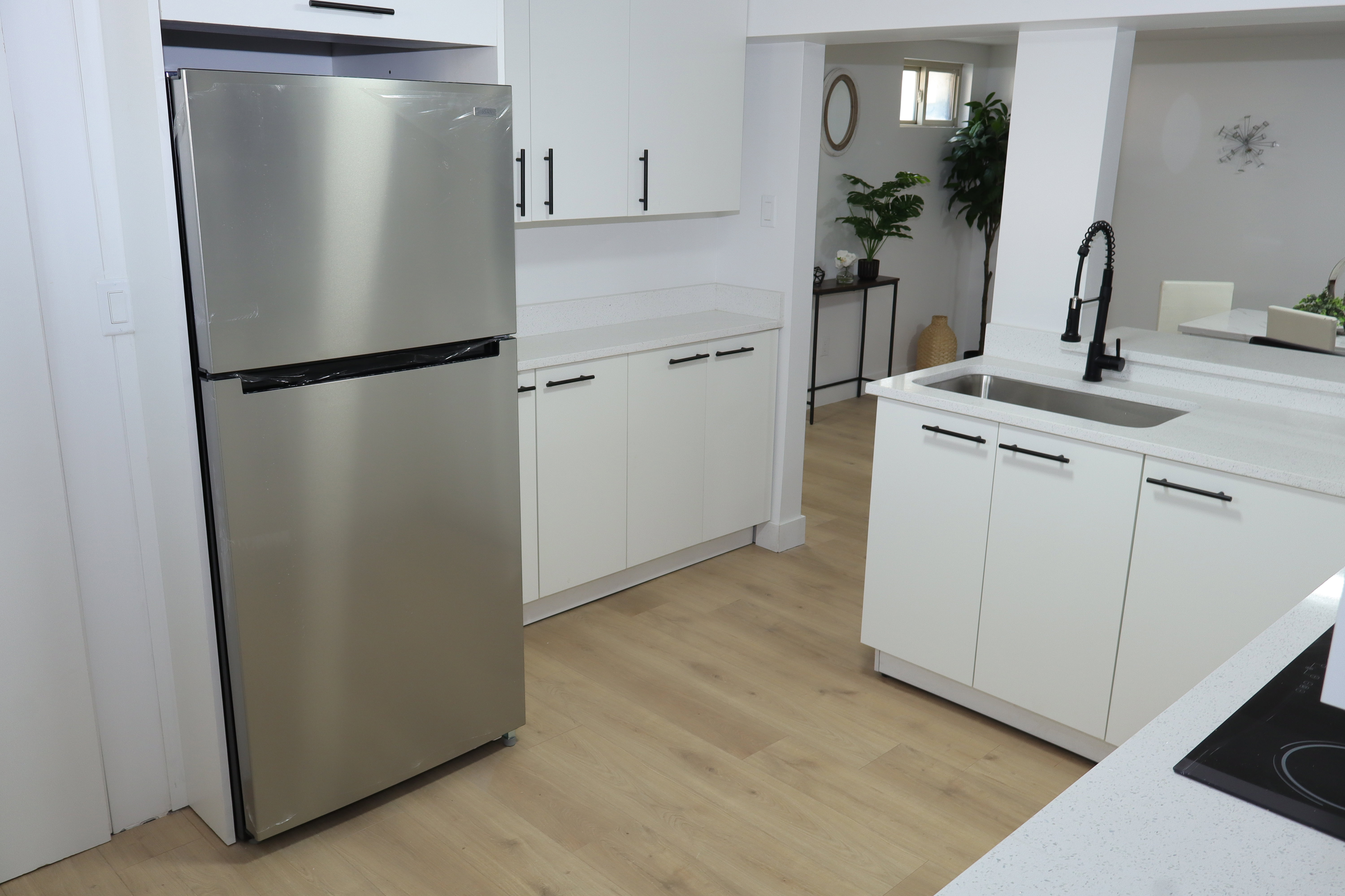 Modern kitchen featuring light wood-finish flooring, white cabinetry with matte black hardware, light-colored countertops, a stainless steel refrigerator, and a kitchen island with an undermount sink and matte black pull-down faucet