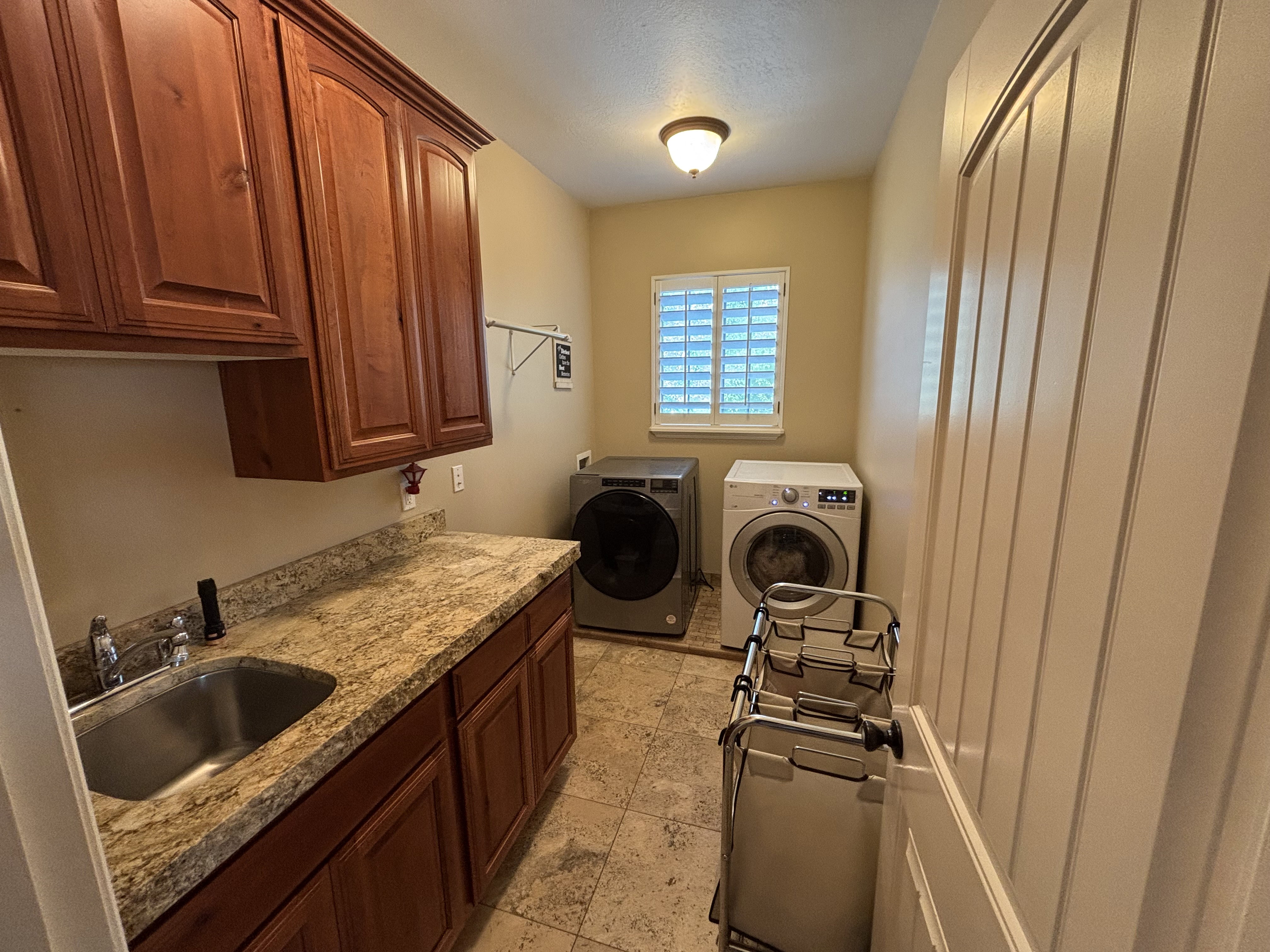Dedicated laundry room featuring warm wood cabinetry, granite-style countertops, a stainless steel utility sink, tiled flooring, and a window with plantation shutters