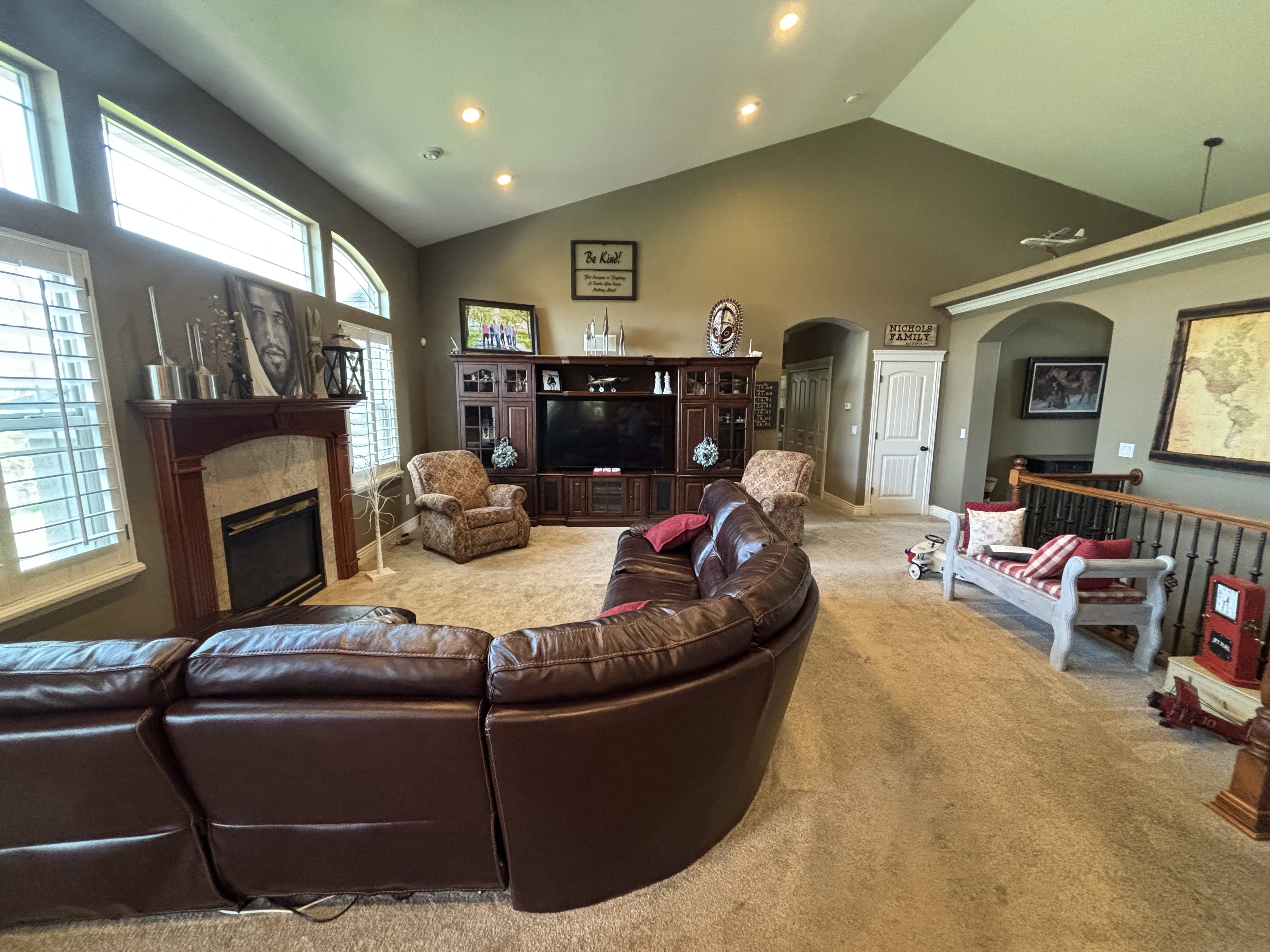Vaulted ceilings with recessed lighting, large windows with plantation shutters, a fireplace with a stone surround and wood mantel, an arched doorway, and a carpeted floor
