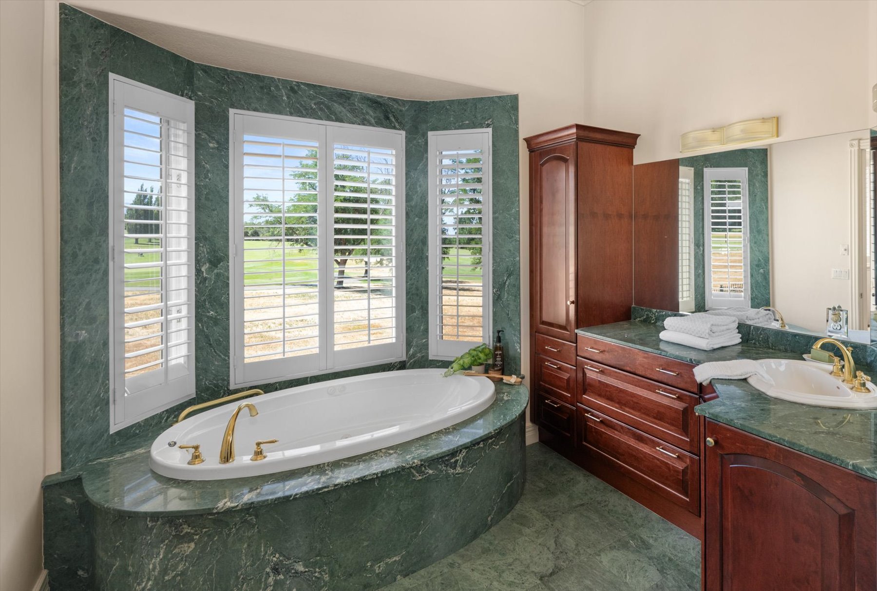 Soaking tub set in a green stone surround with brass fixtures and plantation shutters
