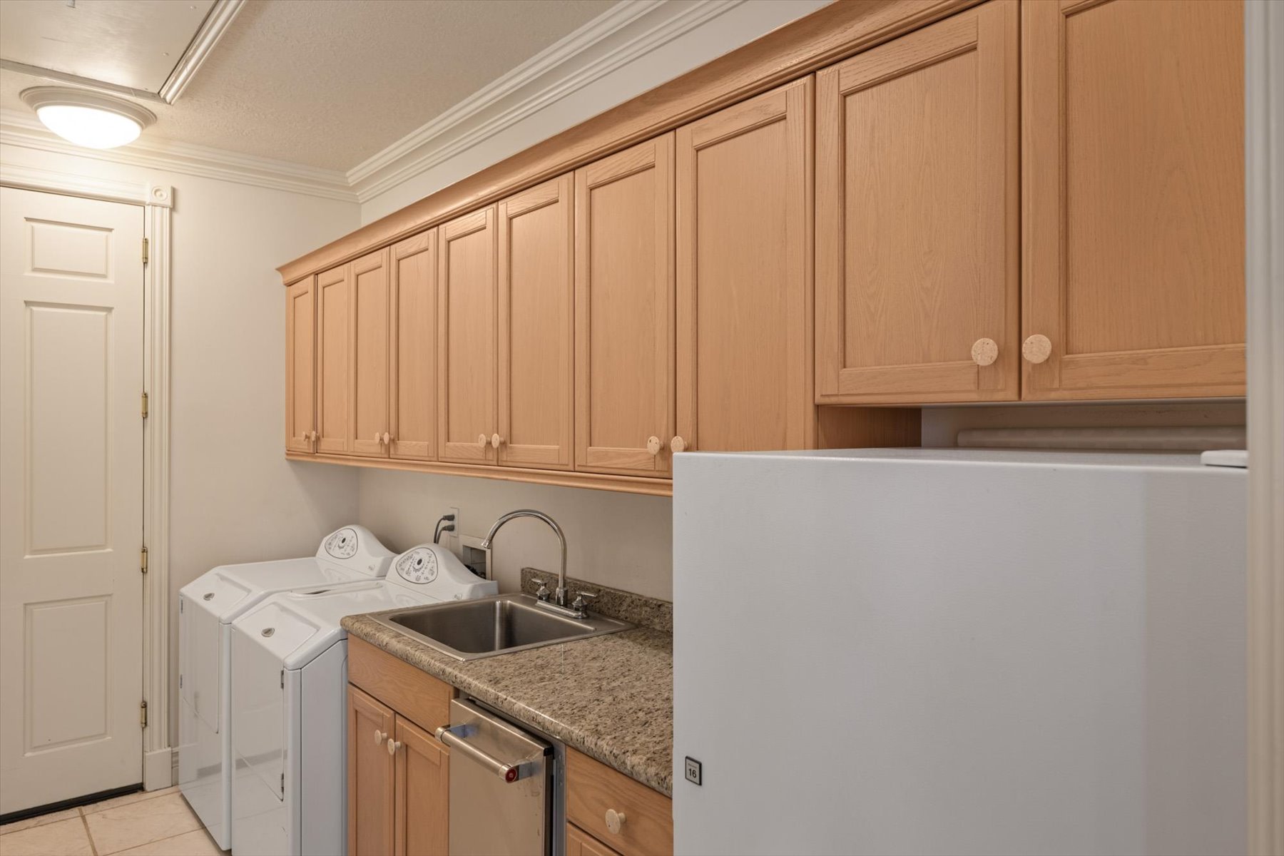 Laundry area with overhead wood cabinetry, granite countertop, and stainless-steel sink with a high-arc faucet, and extra refrigerator off kitchen
