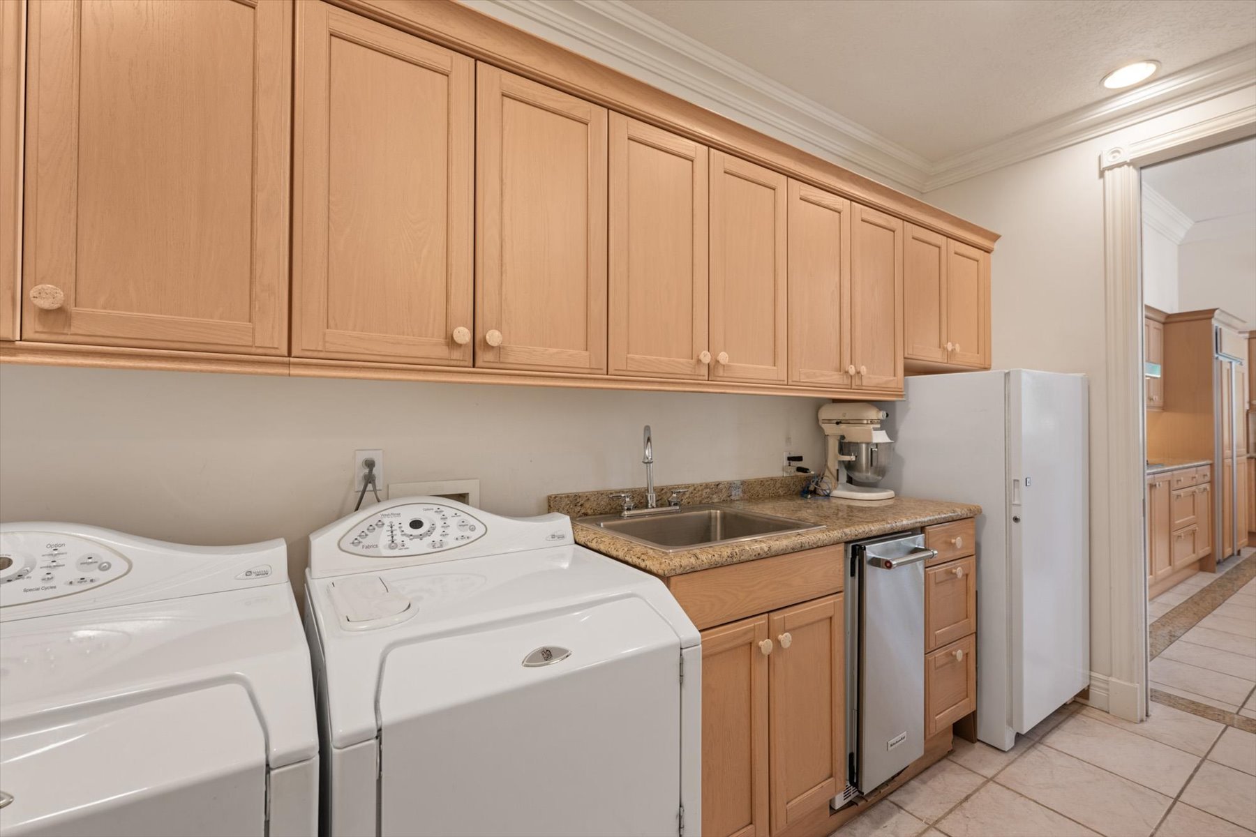 Utility space featuring extensive upper and lower cabinetry, a dual-basin sink with a gooseneck faucet, light-toned countertop, and tiled flooring, between kitchen and garage