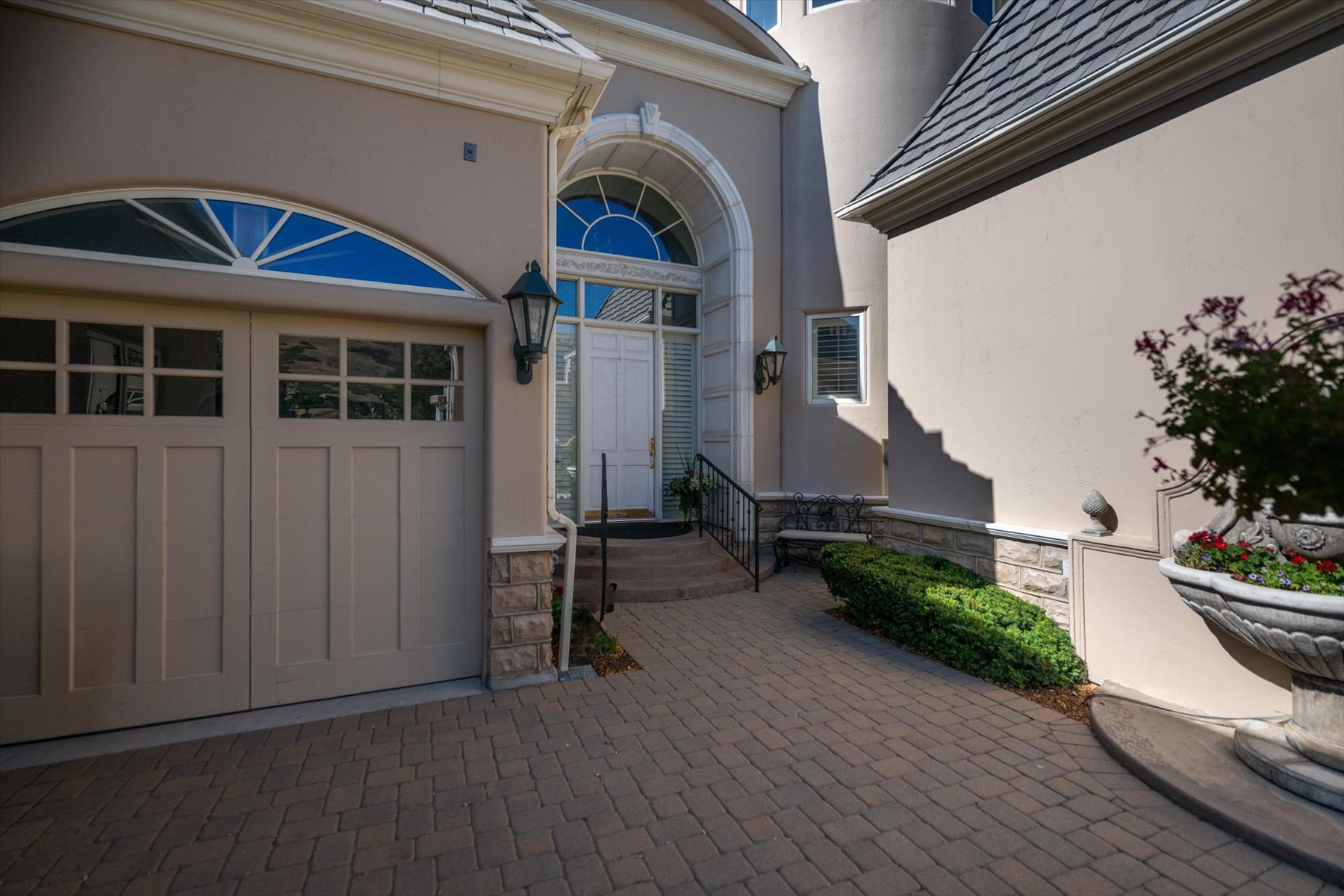 Paver driveway leading to a two-car garage with arched transom window, a grand entry door with sidelights and transom, exterior sconce lighting, and stone veneer accents