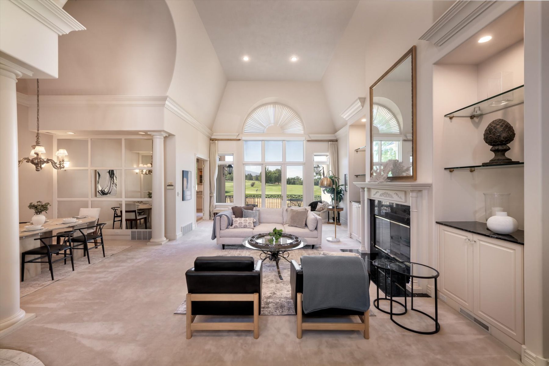 Spacious living area featuring vaulted ceilings, a large arched window, a fireplace with a stone mantel, built-in cabinetry with glass shelving, and light-toned carpeting