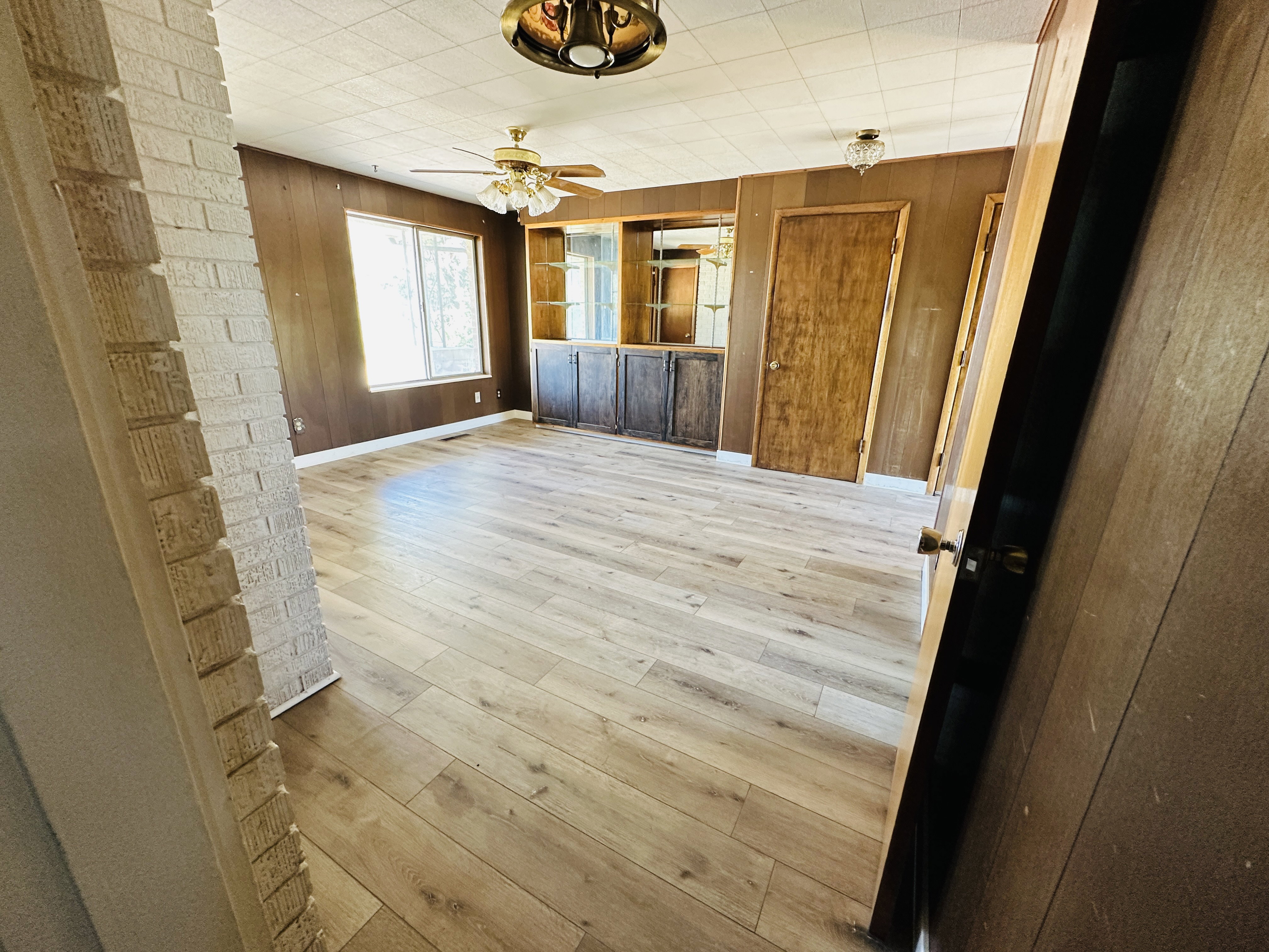 Spacious room featuring wood-finish flooring, wood paneling, a large window, and built-in cabinetry with glass display shelves