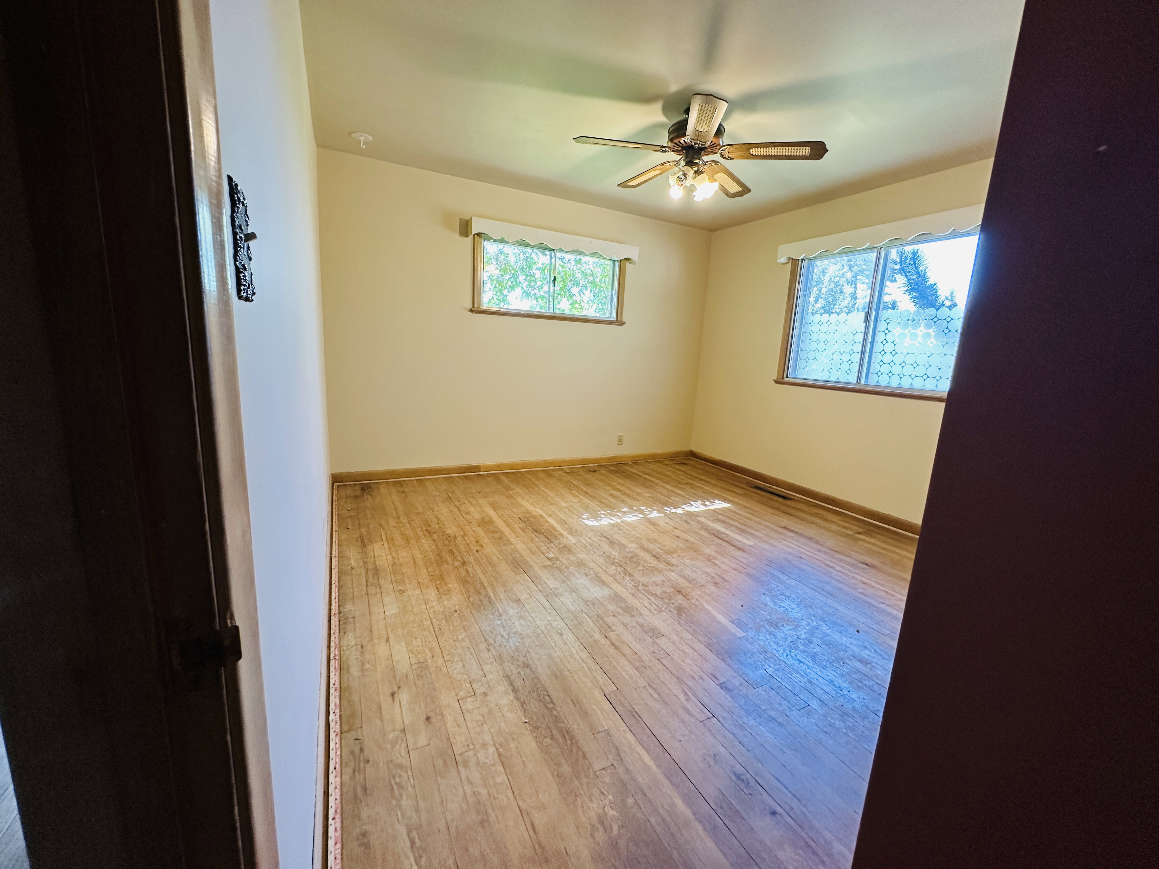 Spacious interior featuring wood-finish flooring, two windows with natural wood frames, a ceiling fan with integrated lighting, and neutral wall tones