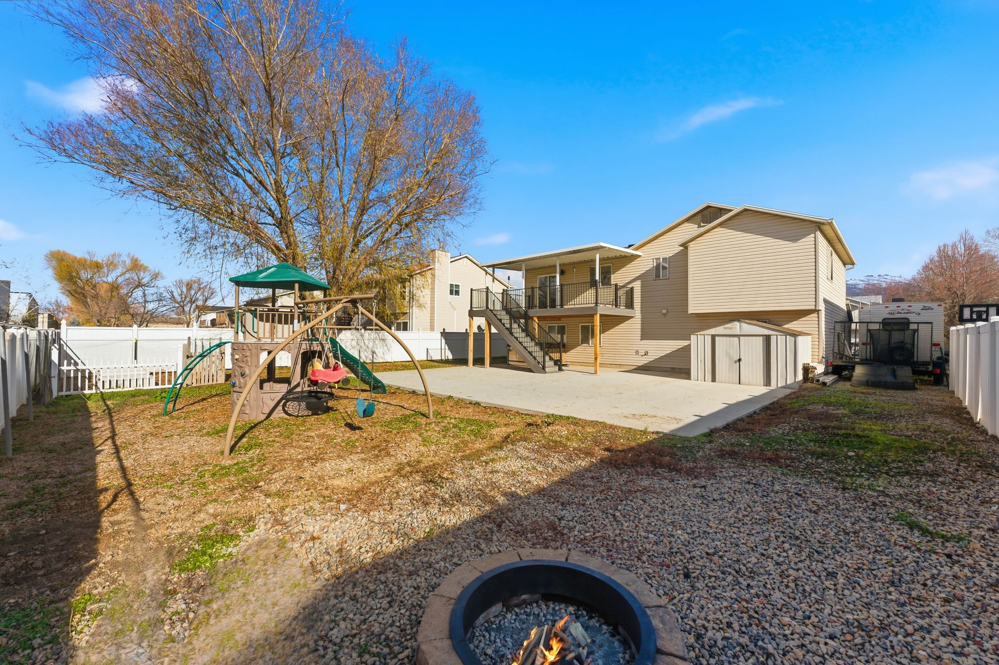 Expansive backyard featuring a large concrete patio, a stone fire pit, and a tan vinyl-sided dwelling with an upper-level deck