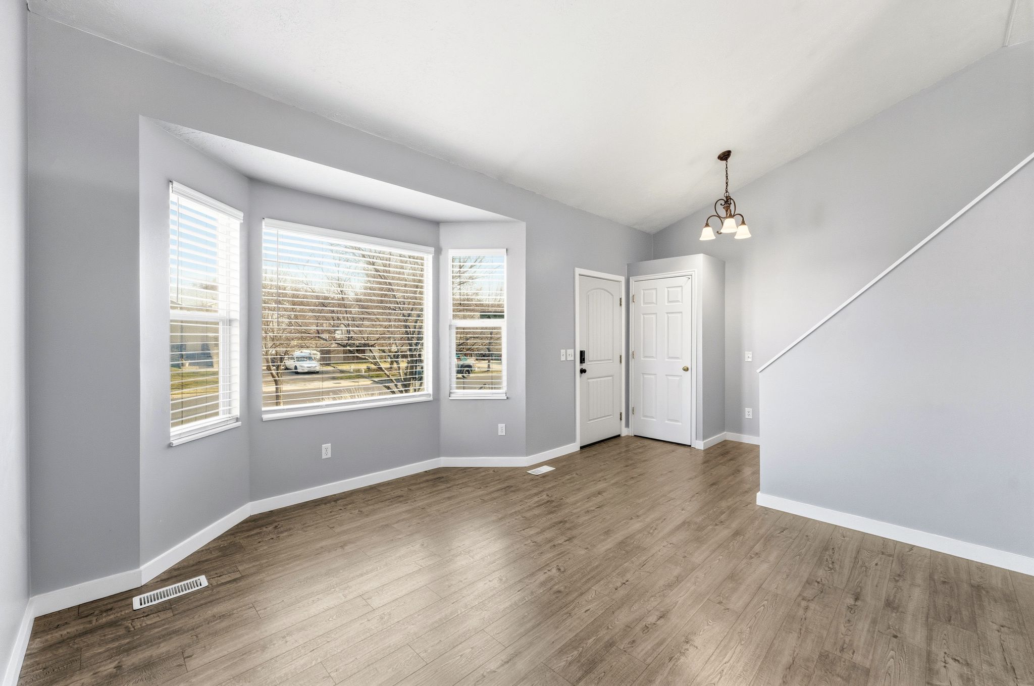 Spacious living area featuring wood-finish flooring, a prominent bay window with blinds, and light gray walls
