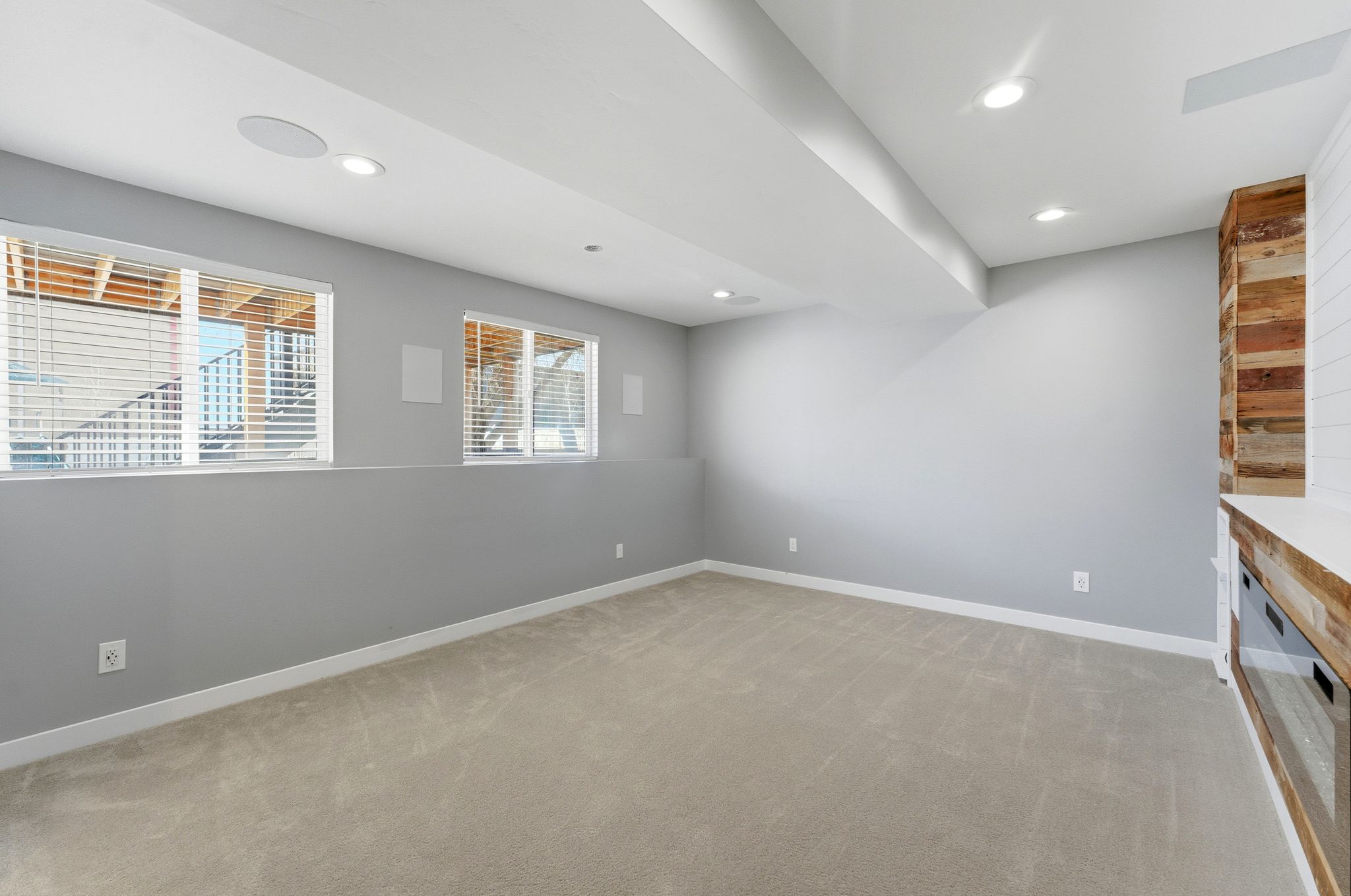Spacious room featuring neutral carpeting, light gray walls, a rustic wood-plank accent wall, recessed lighting, and dual windows with blinds