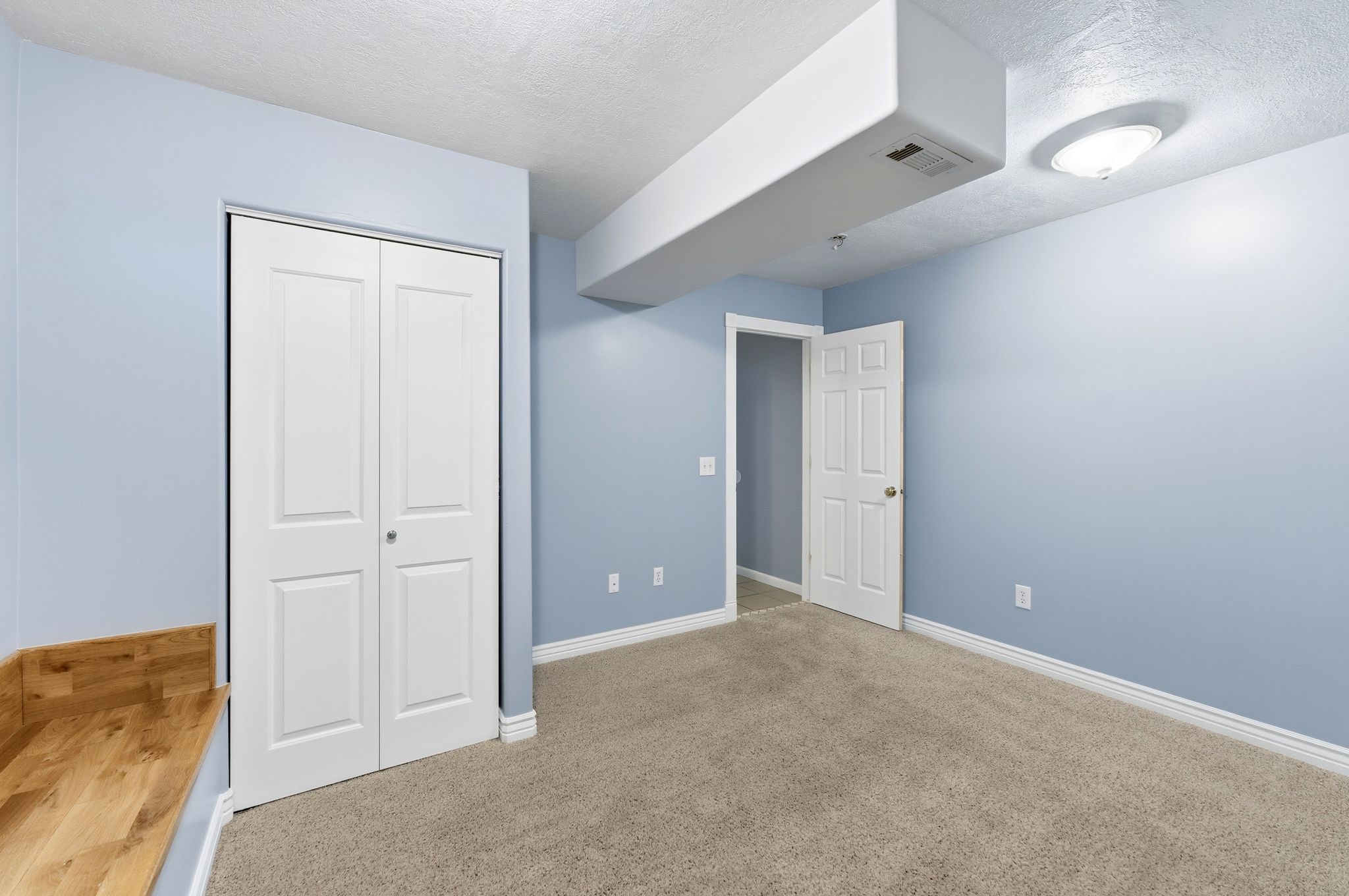 Carpeted room with light blue walls, featuring a white bi-fold closet door, a solid panel interior door, white baseboards, and a wood-finish bench