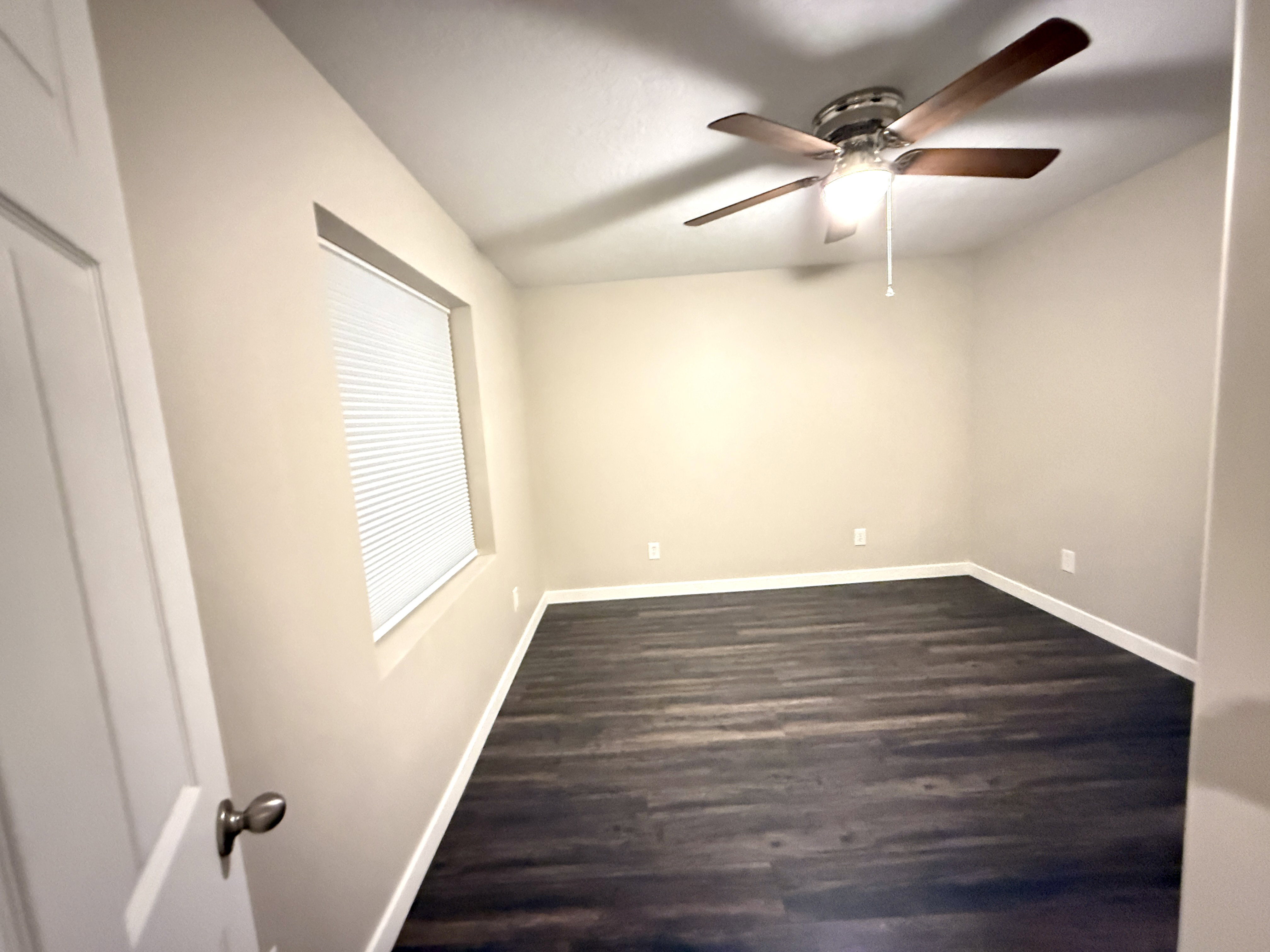 Spacious room featuring wood-finish flooring, a contemporary ceiling fan with integrated lighting, neutral wall tones, and a window with blinds
