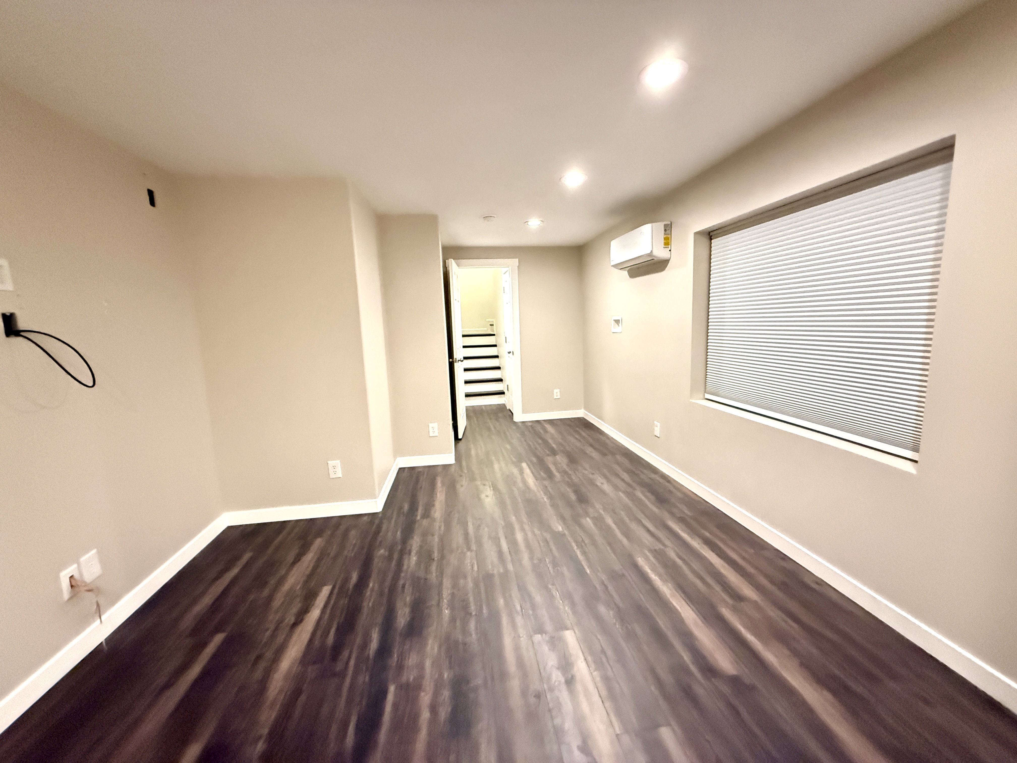 Spacious interior room featuring dark wood-finish flooring, light neutral-toned walls, recessed ceiling lighting, a window with horizontal blinds, and a wall-mounted mini-split AC unit