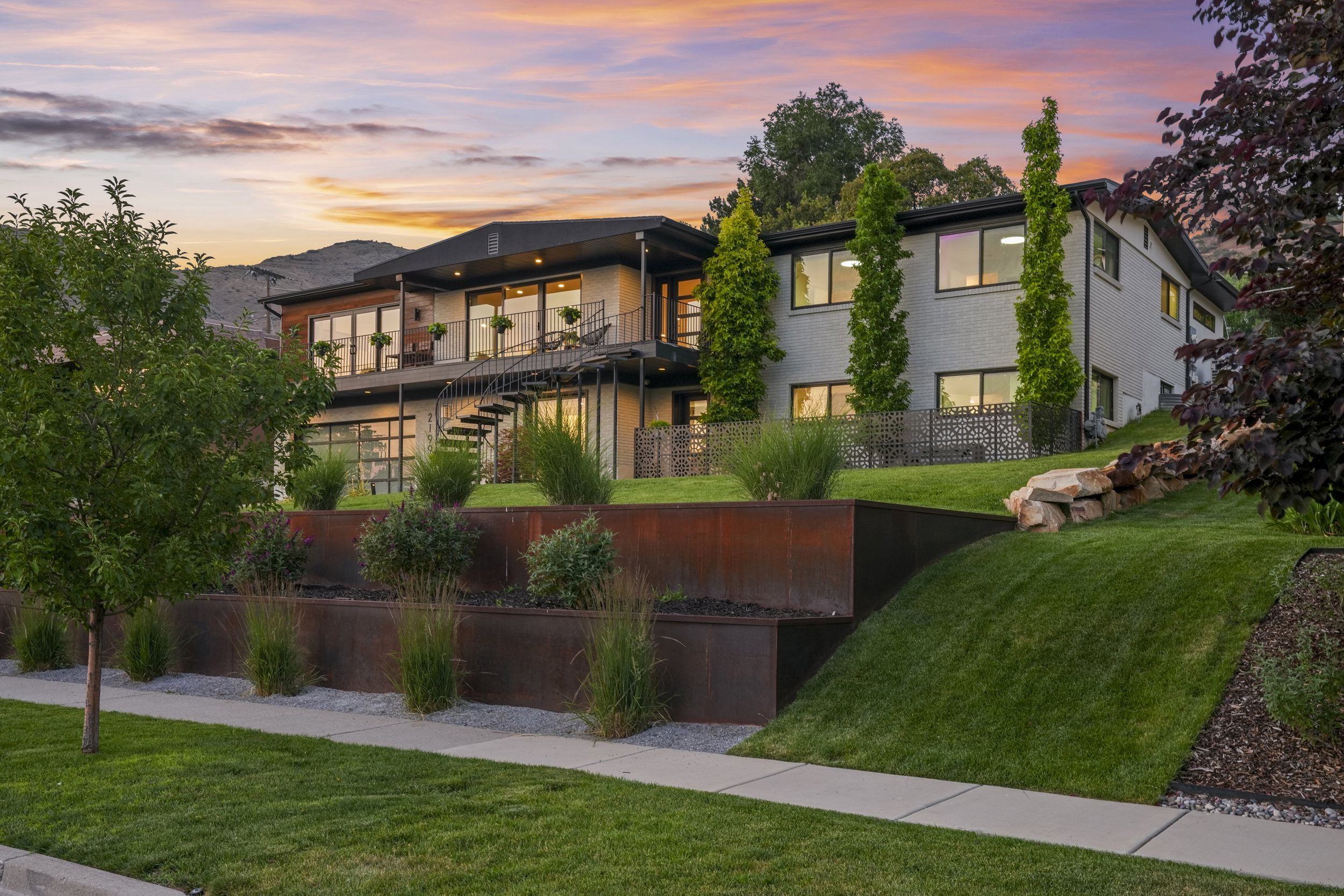 Contemporary multi-level residence featuring expansive windows, a full-width upper balcony with metal railings, and an exterior spiral staircase
