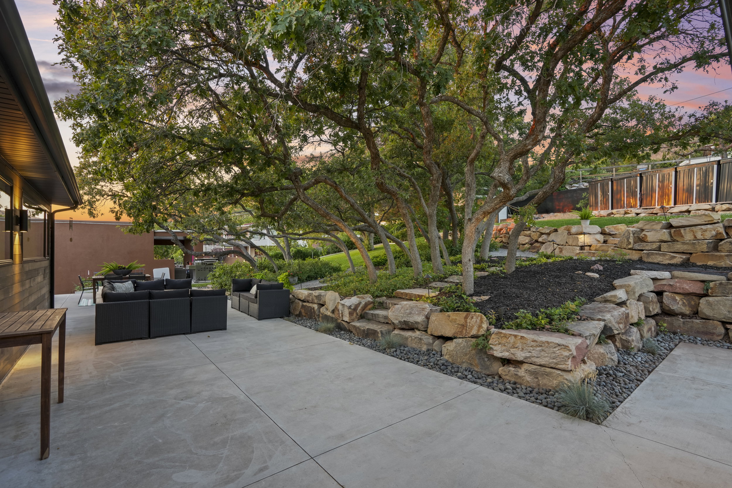 Expansive concrete patio with integrated landscape lighting, mature trees providing natural shade, and tiered stone retaining walls with planted beds