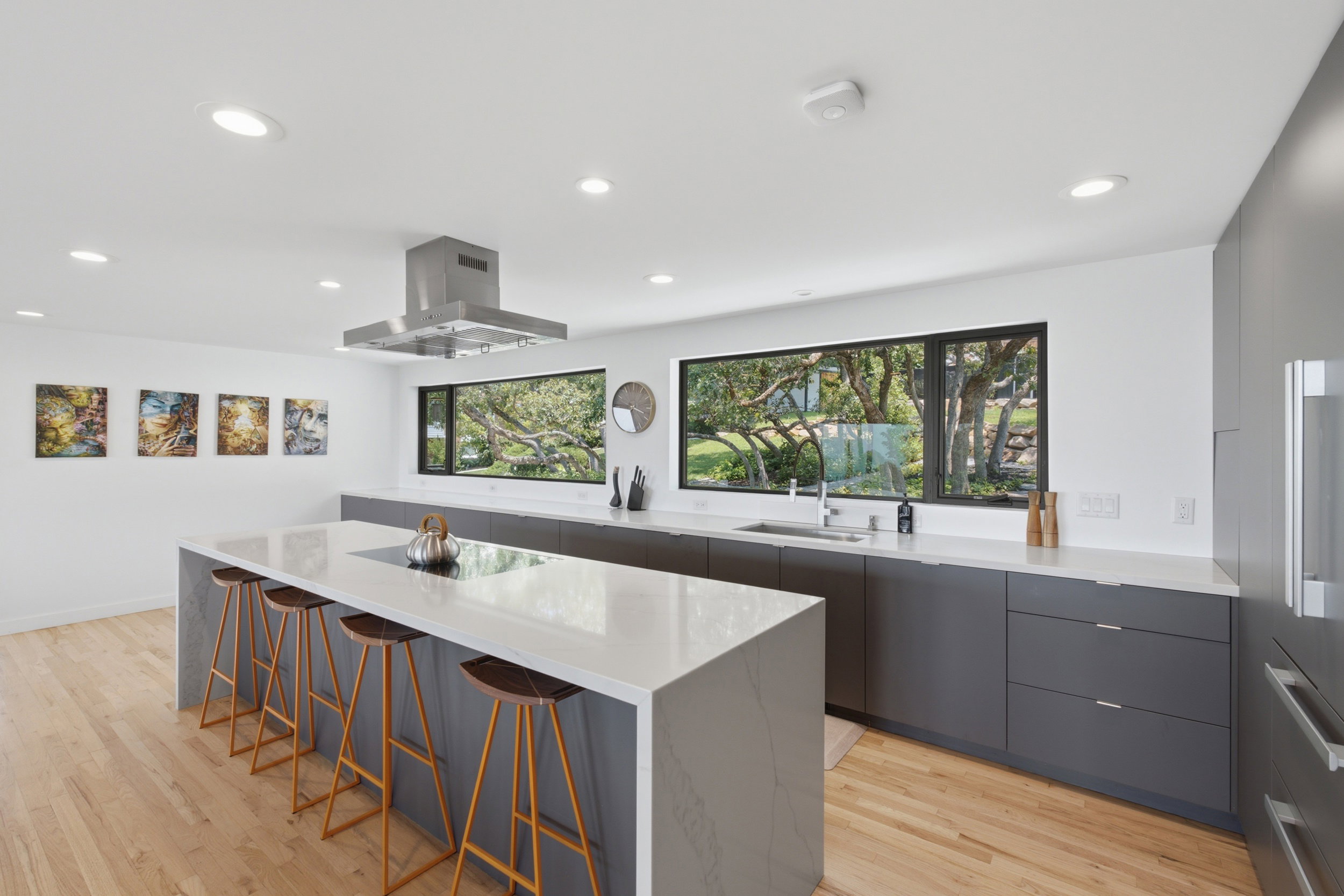 Modern kitchen featuring a large waterfall-edge island with seating, extensive dark gray cabinetry, light wood-finish flooring, recessed lighting, and a stainless steel range hood