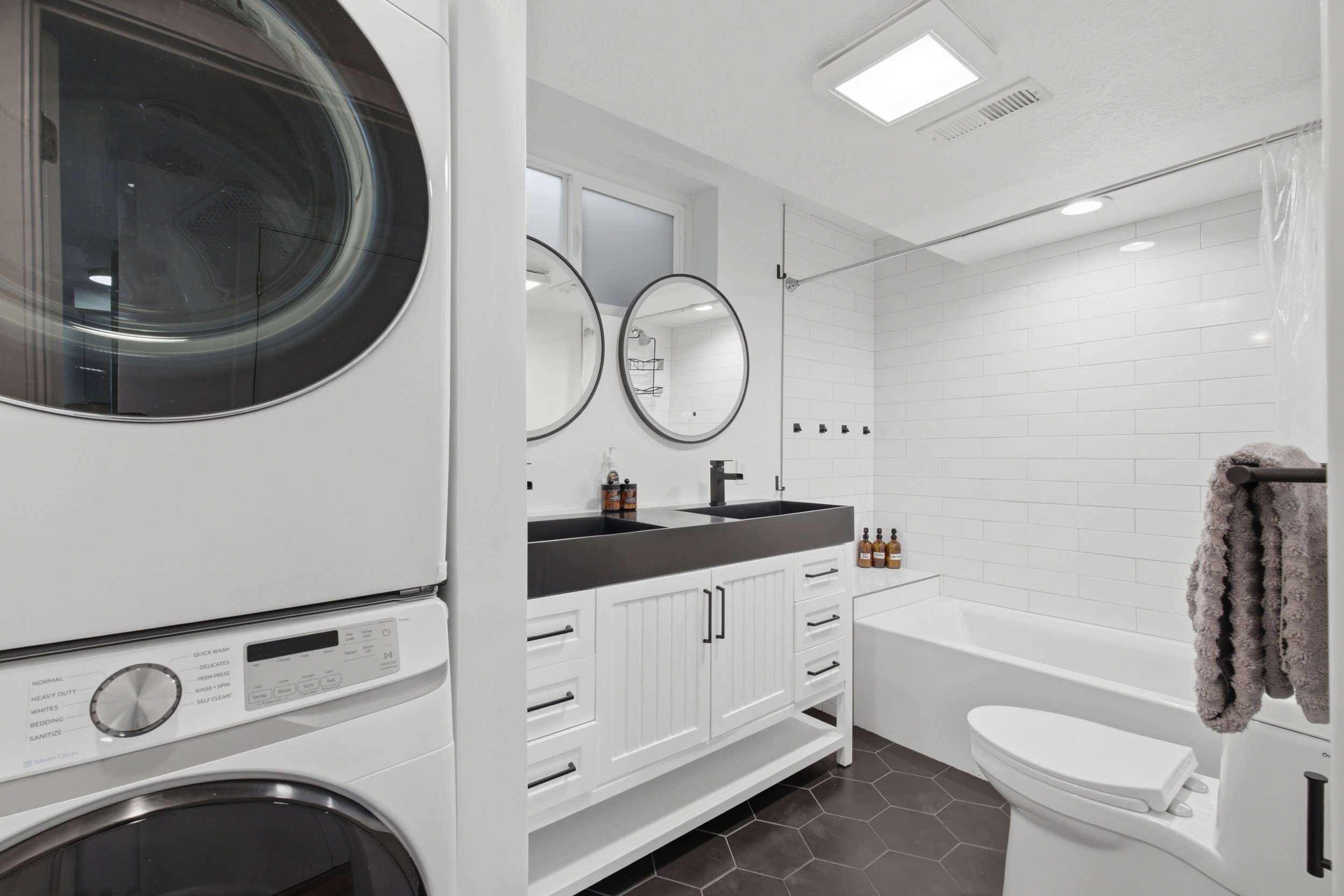Bathroom featuring a stacked washer and dryer unit, double vanity with dual round mirrors, dark hexagonal floor tiling, a white subway tile shower surround, and a white soaking tub