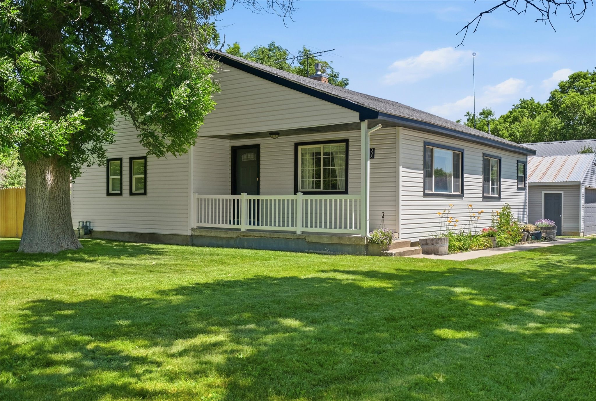 Single-story residence featuring light-colored siding, black trim accents, a covered front porch with white railings, a front entry door with a glass panel, and a side outbuilding