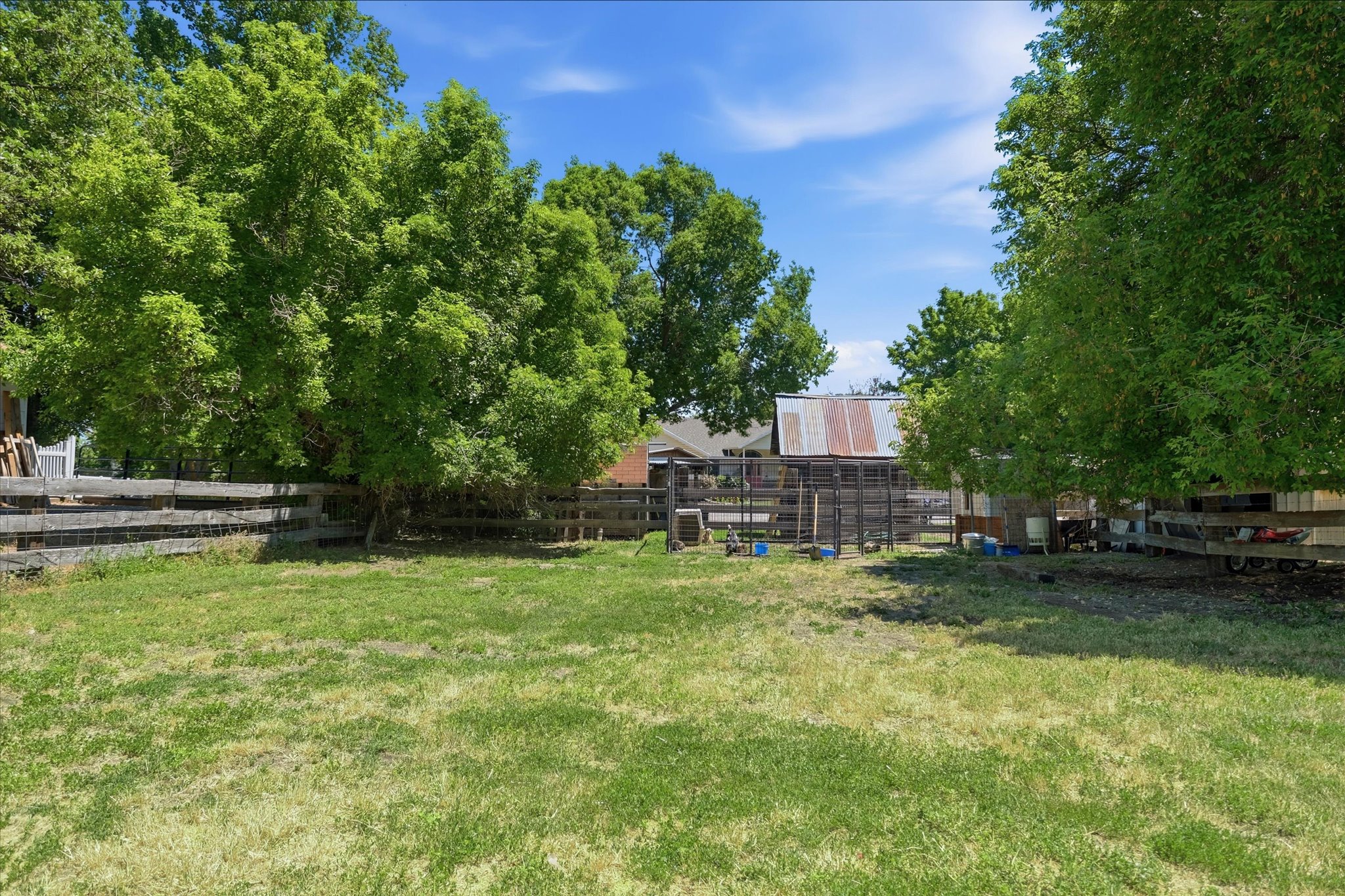 Expansive grassy yard with a wooden fence boundary, mature trees providing natural shade, and outbuildings featuring a corrugated metal roof