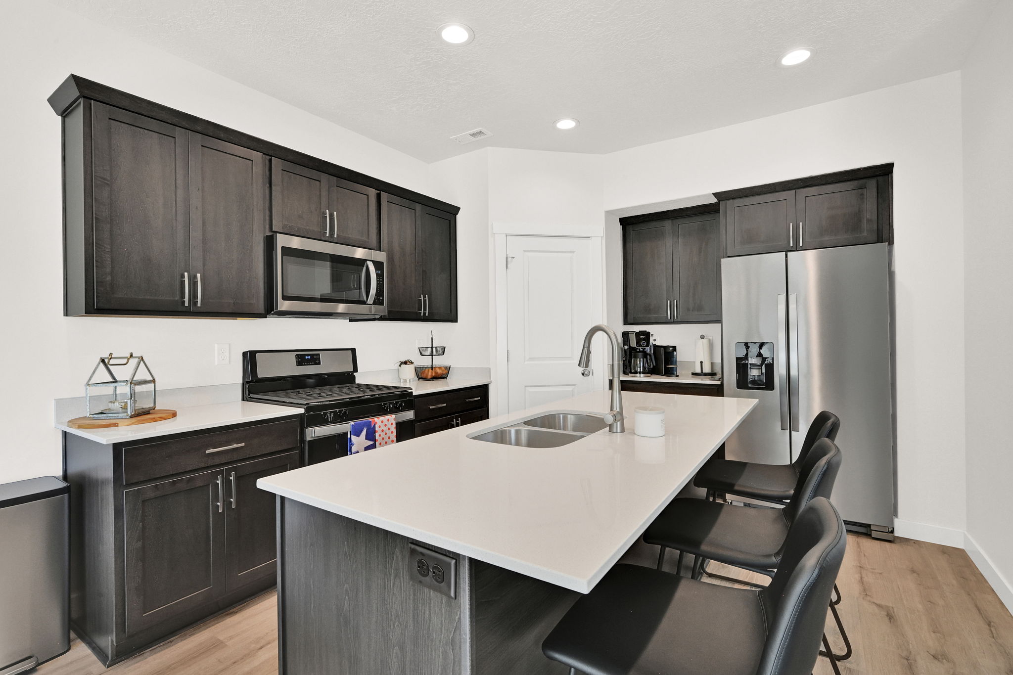 Contemporary kitchen featuring dark wood cabinetry, stainless steel appliances, a center island with double sink, light-toned countertops, and recessed lighting