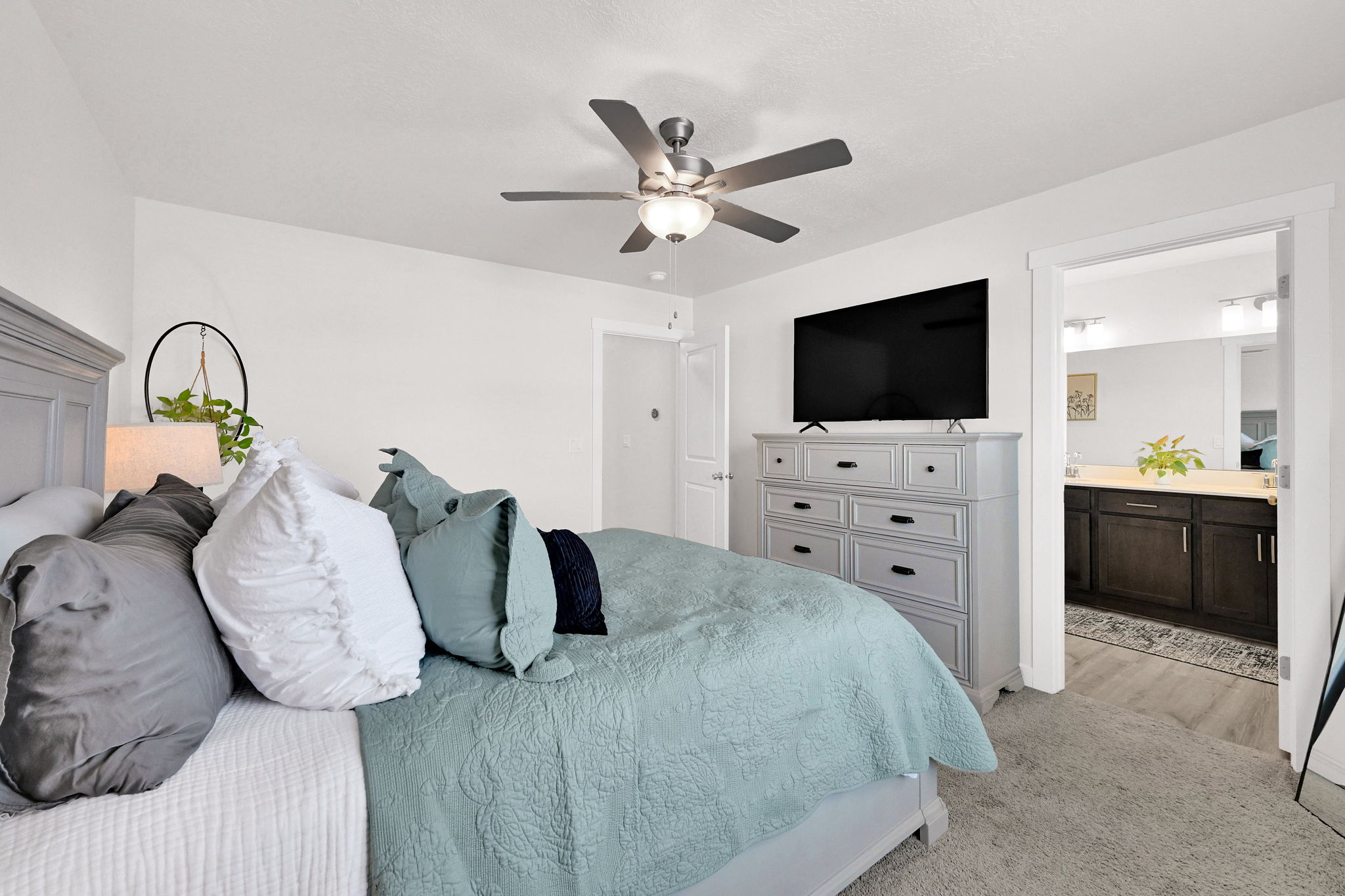 Carpeted room featuring a ceiling fan with integrated lighting, white walls, and a substantial doorway revealing a bathroom with a dual vanity, light-colored countertop, and dark wood-finish cabinetry