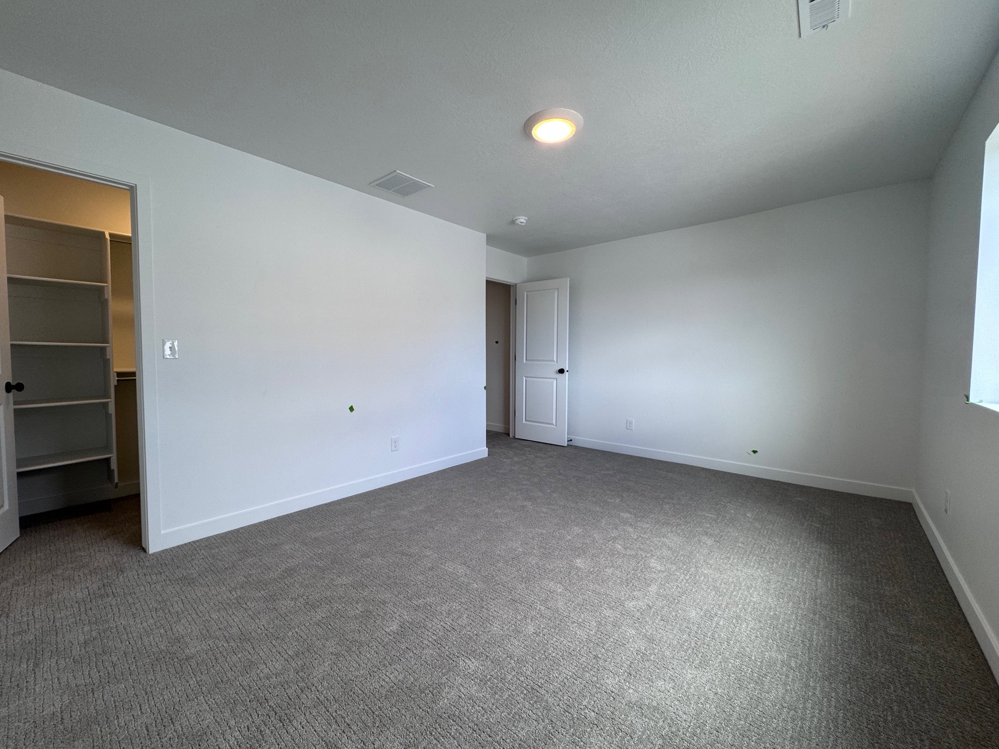 Spacious room featuring neutral carpeting, white painted walls, a built-in closet with shelving, a white door with black hardware, and a ceiling-mounted light fixture